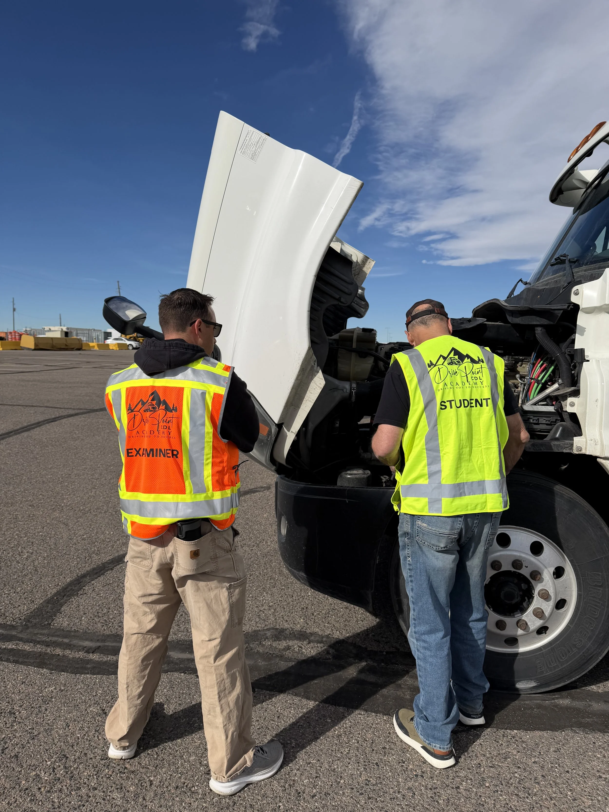 Two automotive students and an examiner working on a large truck engine in an outdoor parking lot under a blue sky.