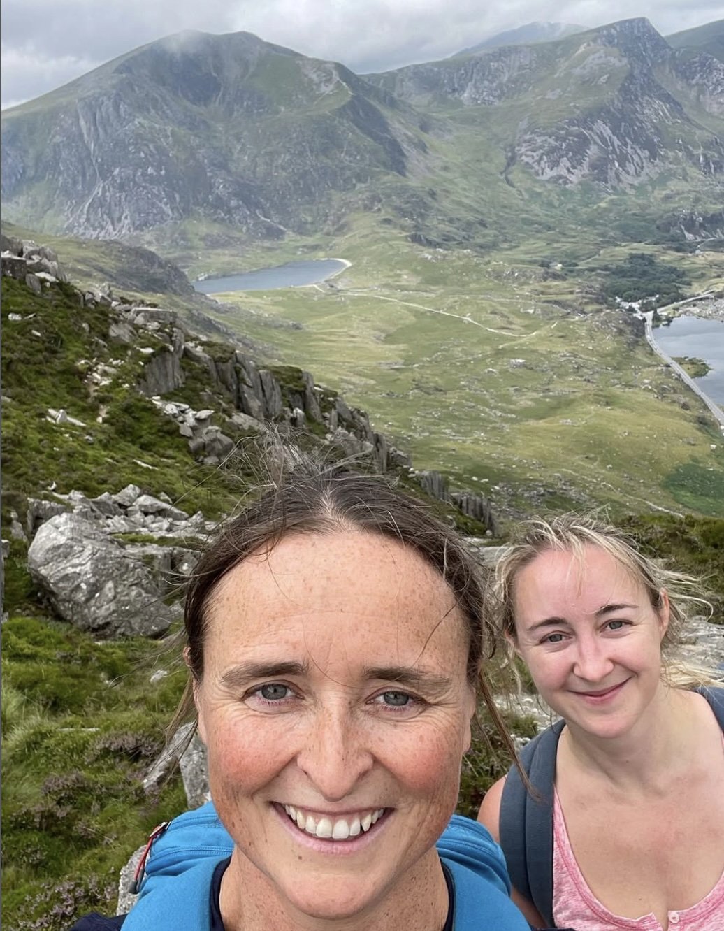 Visit Wild Wales with Emily Coates smiling outdoors in a mountainous landscape with lakes, rocky terrain, and green hills.