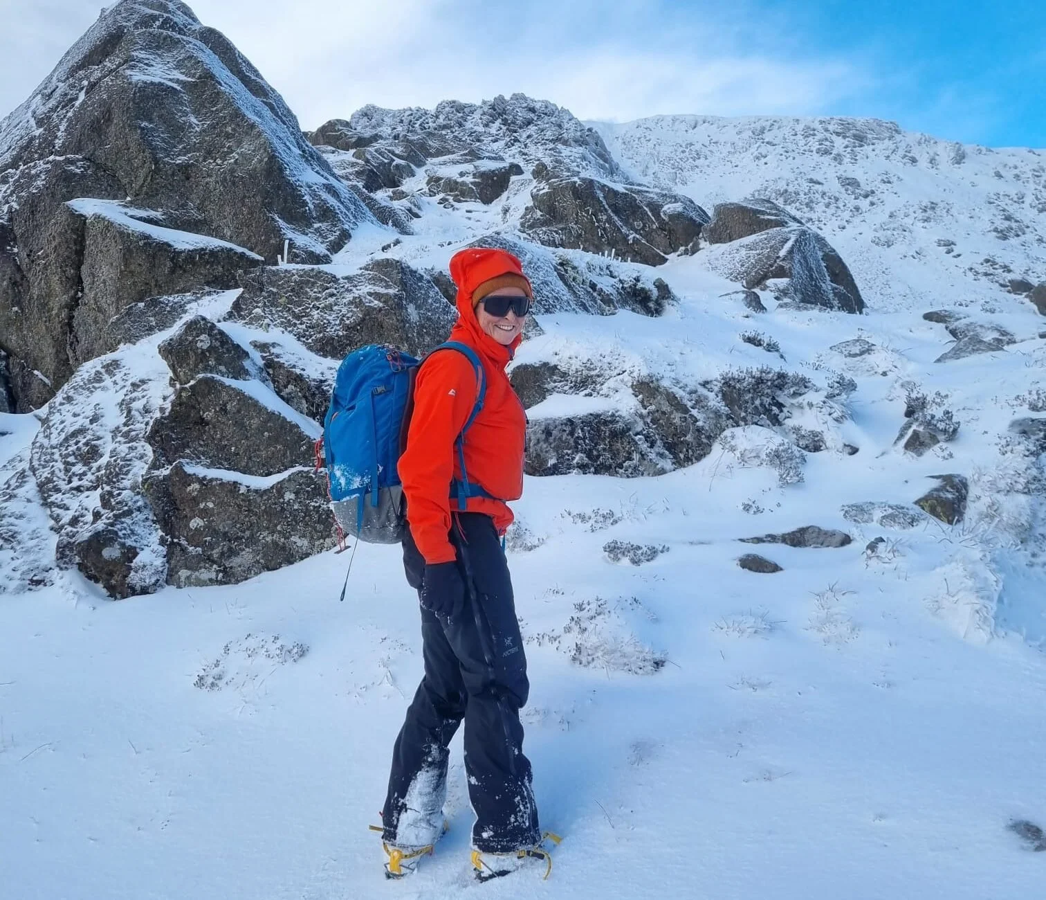 Visit Wild Wales with Emily Coates. Person in red jacket and snow boots standing in snow-covered mountain landscape with rocks and snow on the ground and mountain peaks in the background.