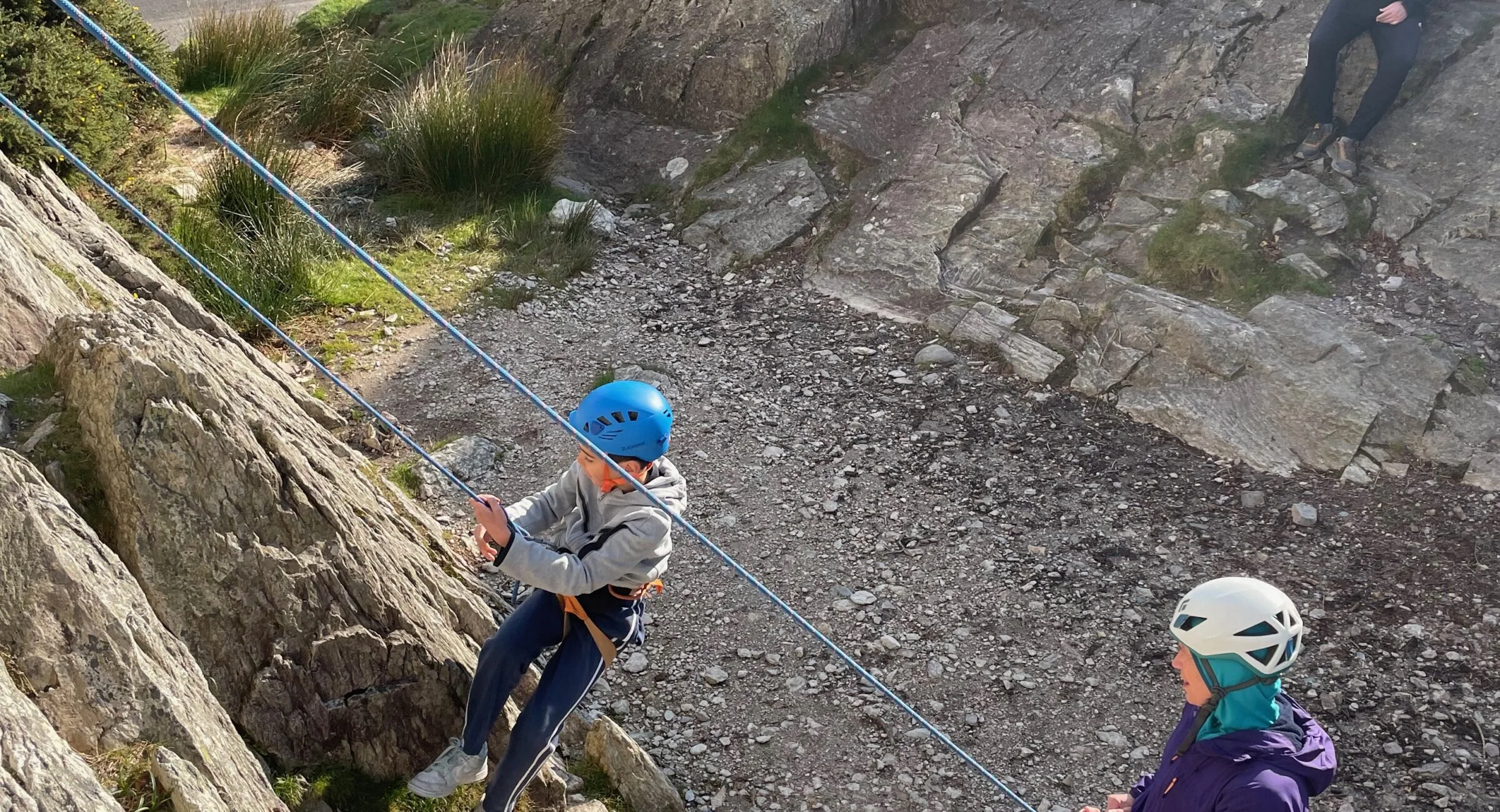 Visit Wild Wales with Emily Coates. Children rock climbing outdoors, wearing helmets and harnesses, holding onto climbing ropes, on rocky terrain with some vegetation.