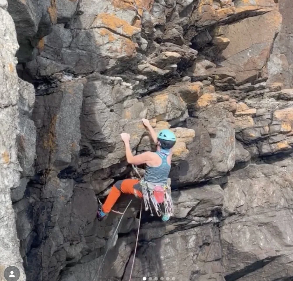 Visit Wild Wales with Emily Coates. A person wearing a blue helmet, striped tank top, and orange pants is rock climbing on a rugged rock wall with climbing gear.