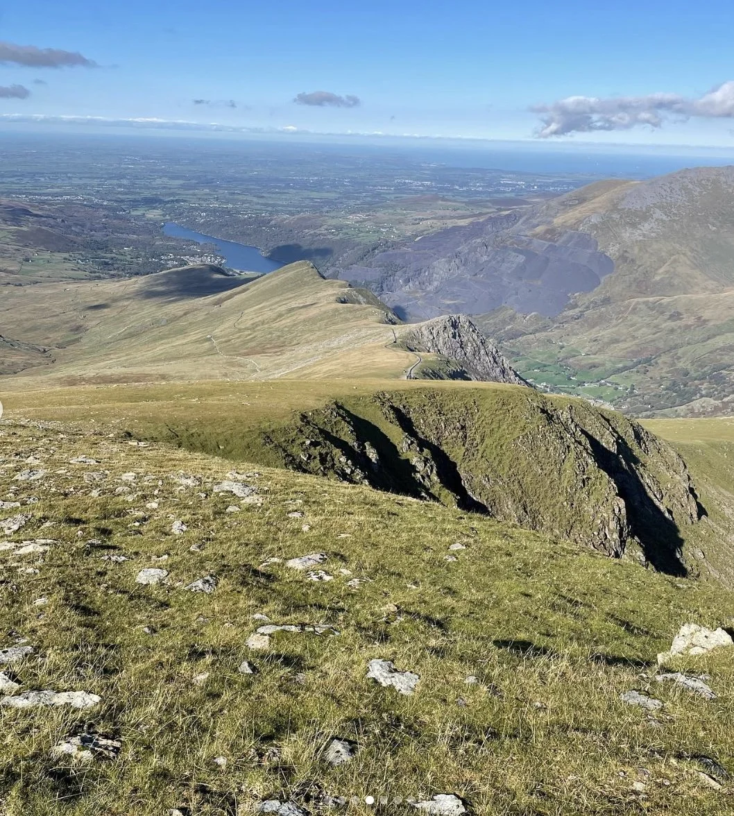 A scenic view from a mountain summit showing rolling grassy hills, rocky cliffs, a distant lake, and a valley with fields and small towns under a blue sky with scattered clouds.