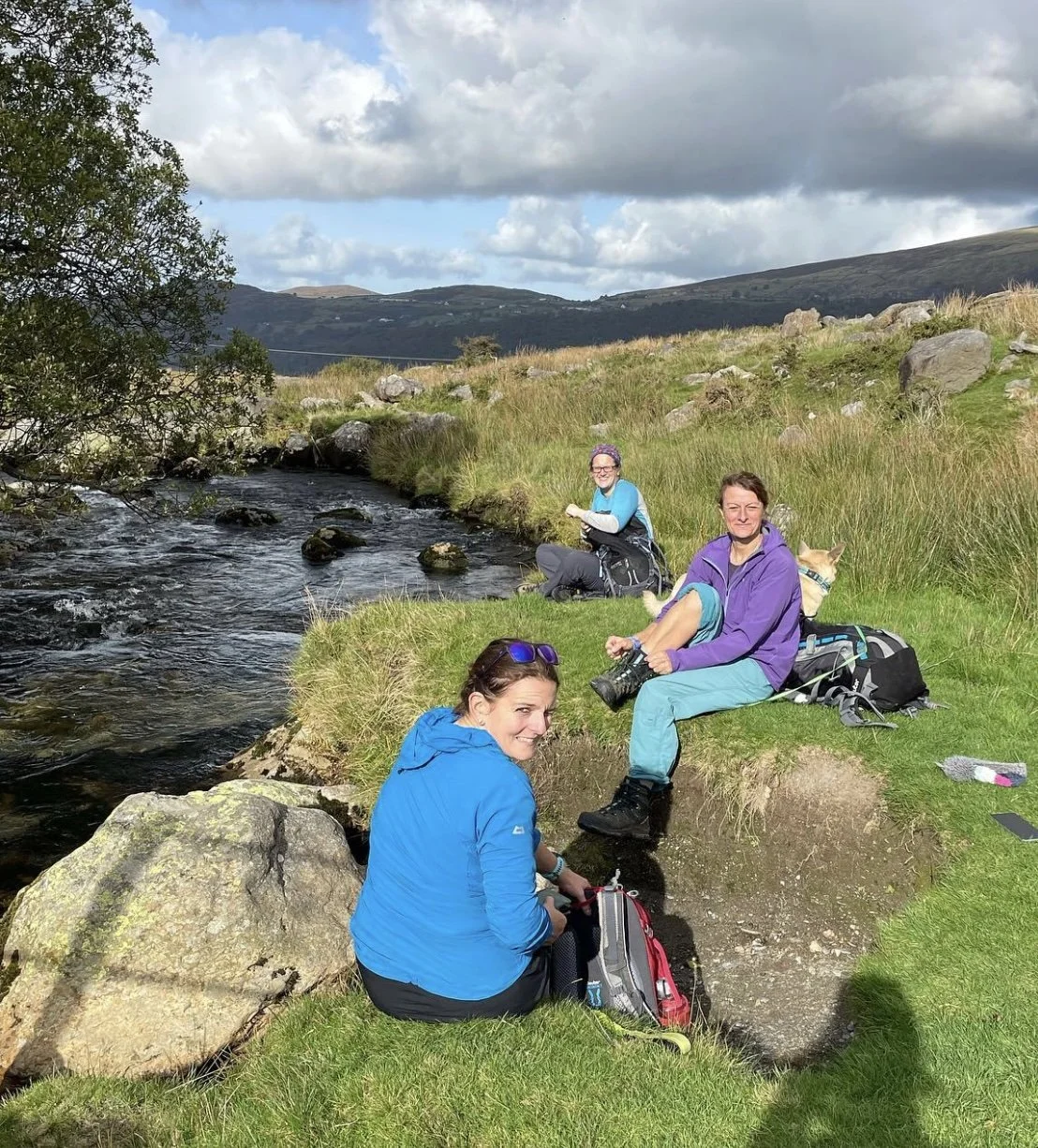 Visit Wild Wales with Emily Coates. Three women with backpacks and a dog sitting near a stream in a grassy landscape with hills and clouds in the sky.
