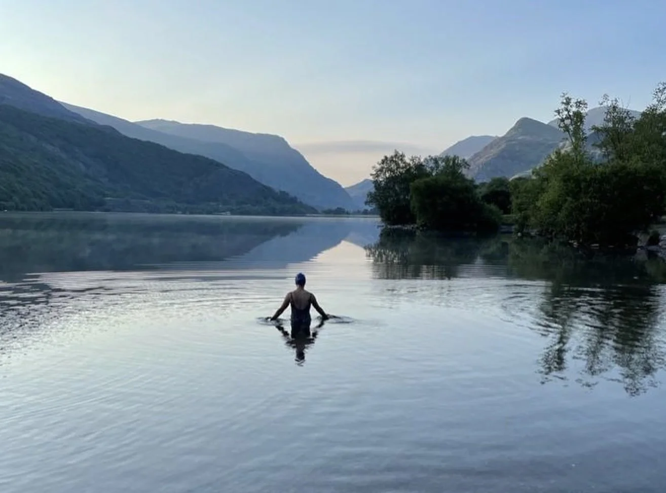 Visit Wild Wales with Emily Coates. A person standing waist-deep in a calm river with mountains in the background during twilight.