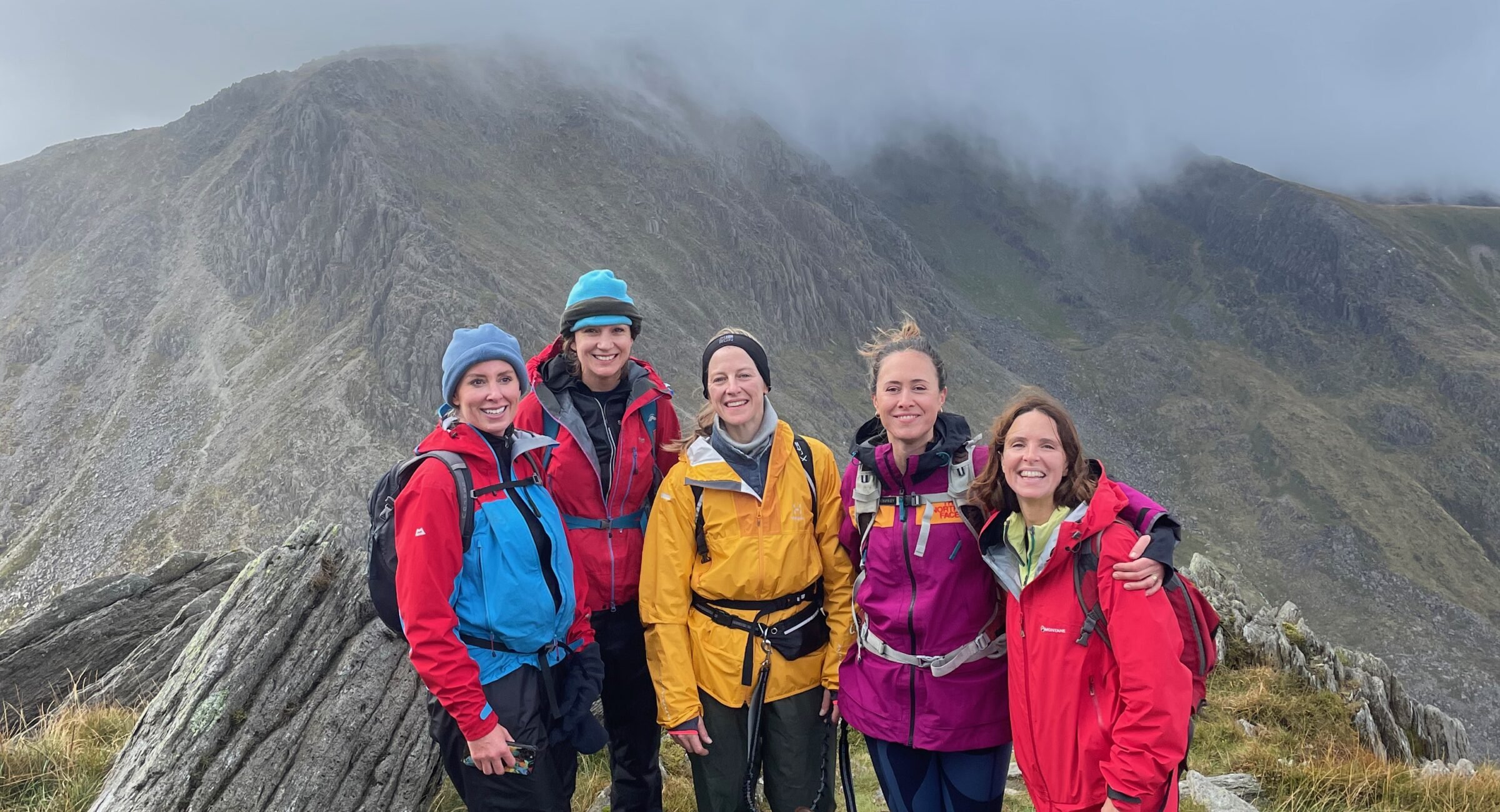 Visit Wild Wales with Emily Coates. Group of five women in colorful outdoor jackets and backpacks standing together on rocky terrain with mountain landscape and foggy sky in the background.