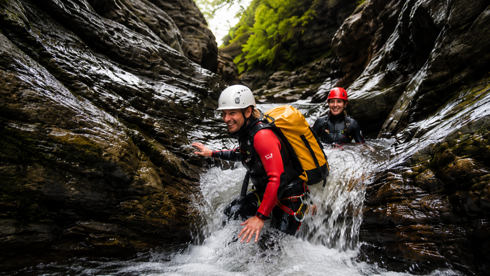 Two people wearing helmets and wetsuits navigating through a narrow canyon with flowing water, surrounded by dark wet rocks and green foliage in the background.