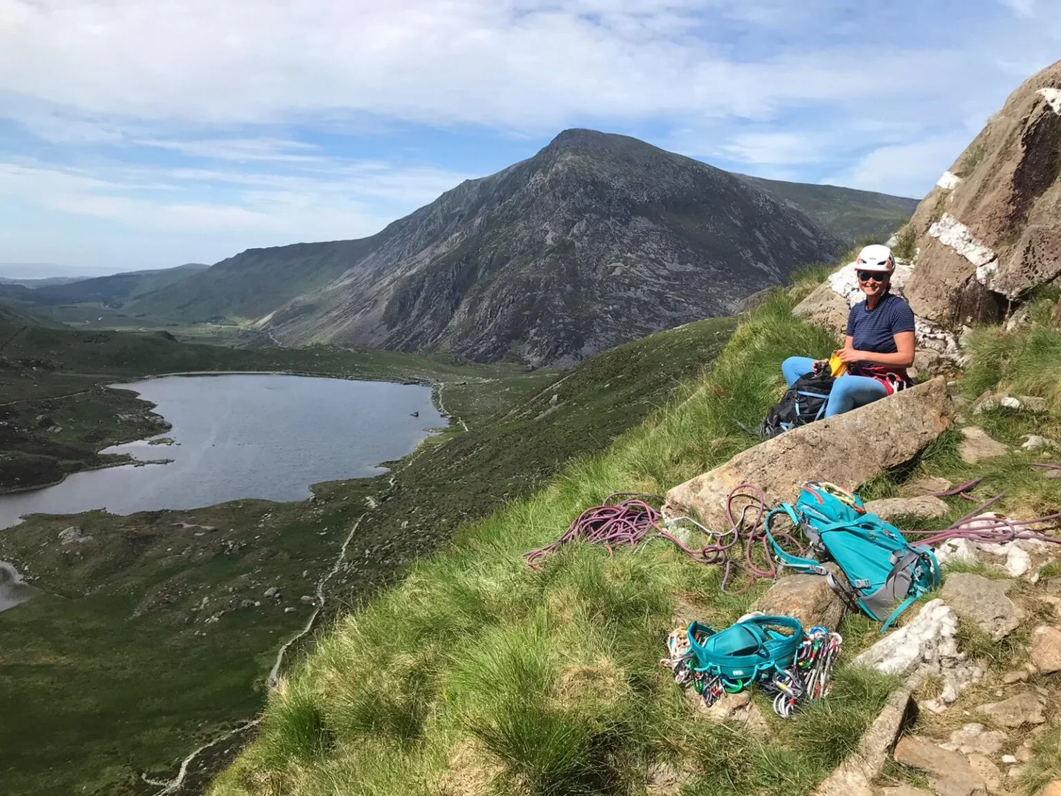 A woman in climbing gear sitting on a rock ledge on a mountain, with climbing ropes, backpacks, and gear around her. She is smiling and wearing a helmet and sunglasses, overlooking a mountain lake and green valley.