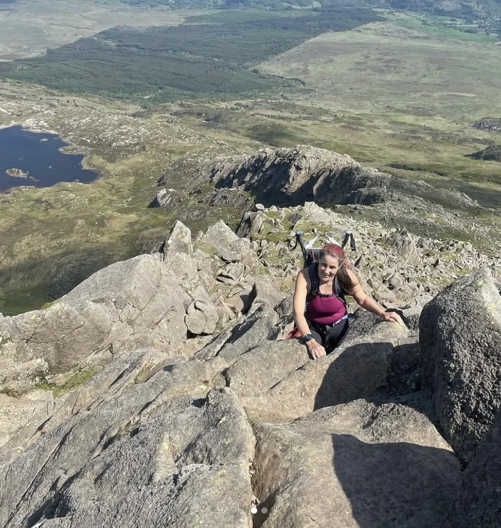 Visit Wild Wales with Emily Coates. A woman climbing a rocky mountain with a landscape of lakes, hills, and fields in the background.