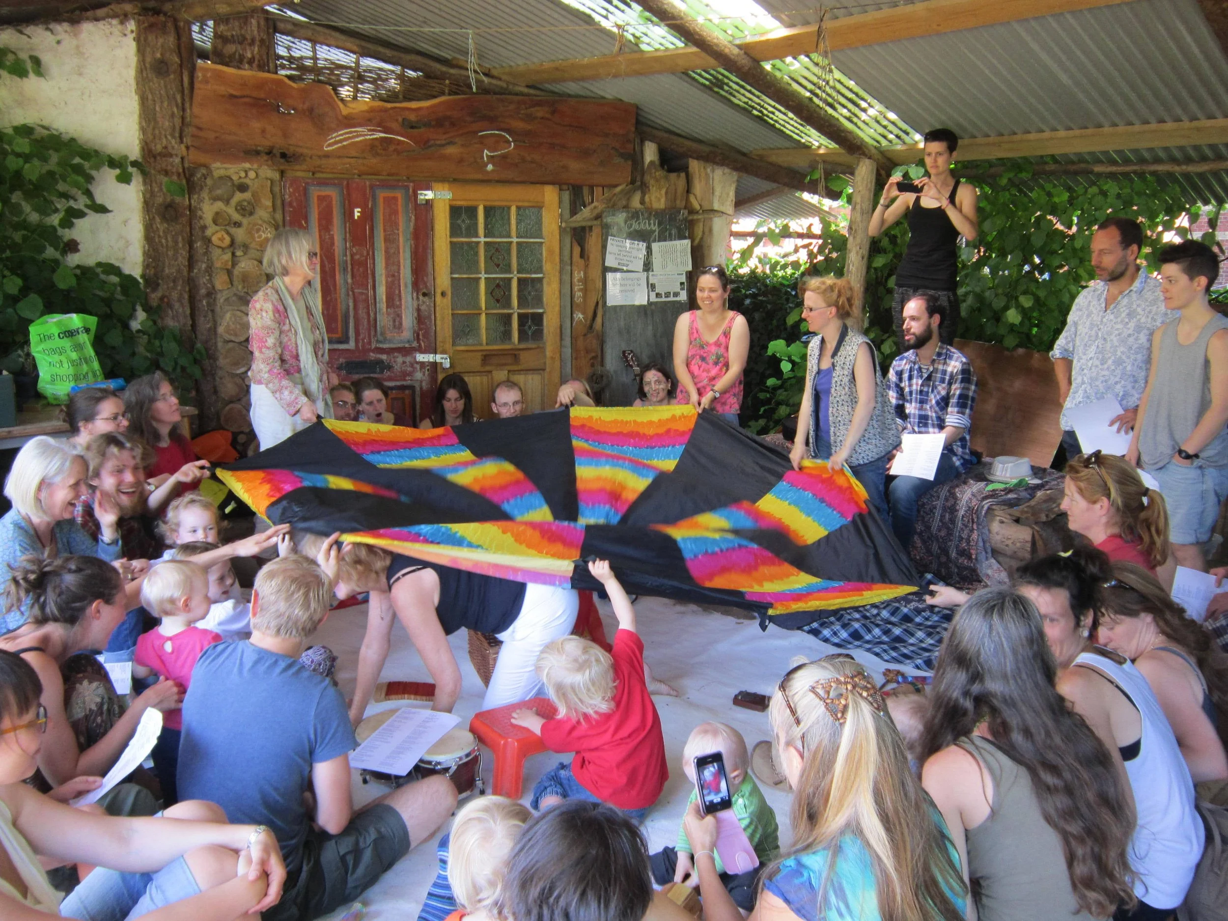 A group of people, including children and adults, gathered indoors under a rustic wooden and metal roof, participating in a colorful activity involving a rainbow tie-dye or fabric. A woman in a pink top and glasses stands, holding up the fabric, while others are seated or standing around, some taking photos or videos, in a lively and cheerful atmosphere.
