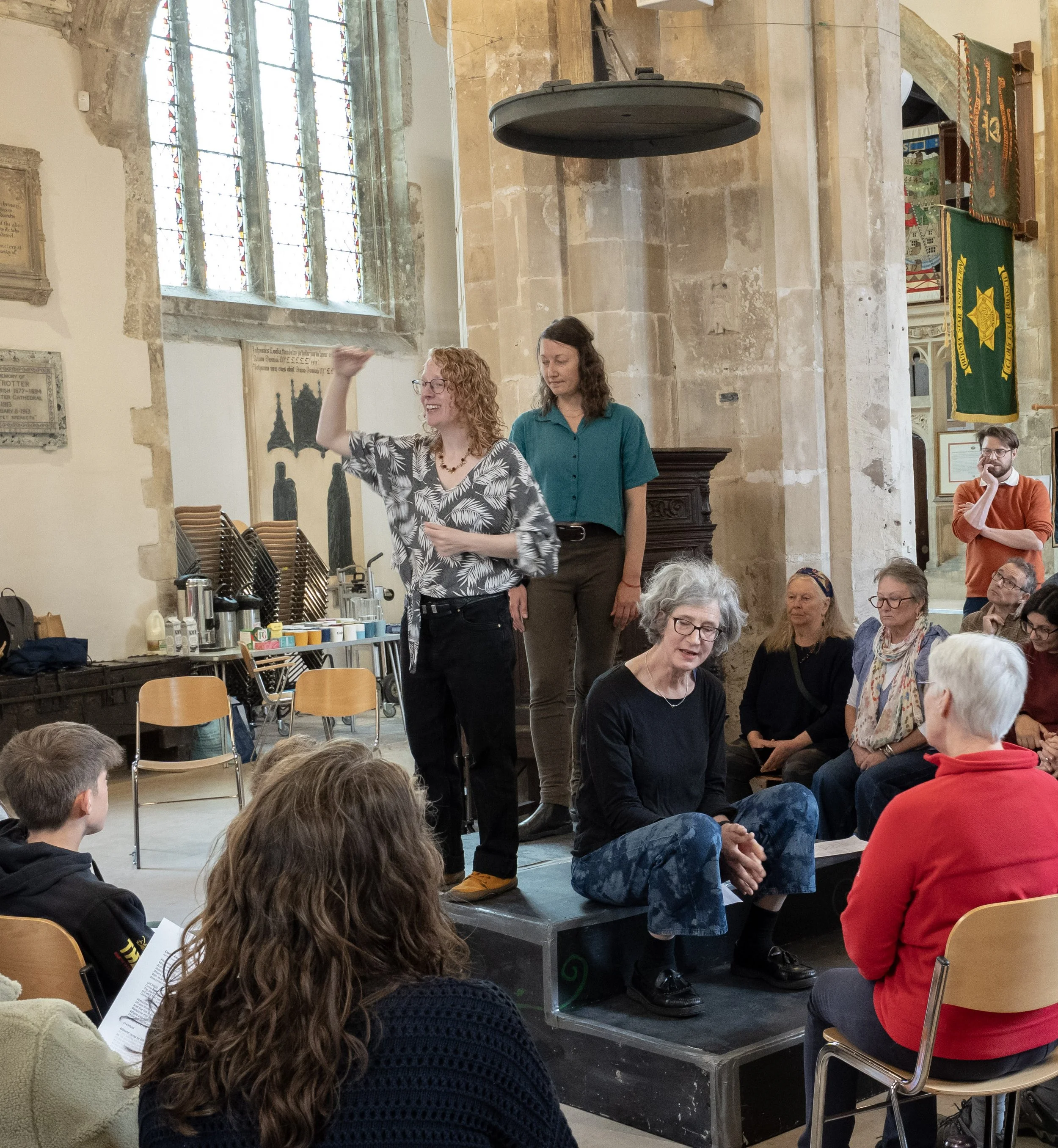 Women and men gathered in a church, with some sitting and some standing, while a woman with curly red hair gestures as she speaks. The room features stained glass windows and banners hanging on the wall.