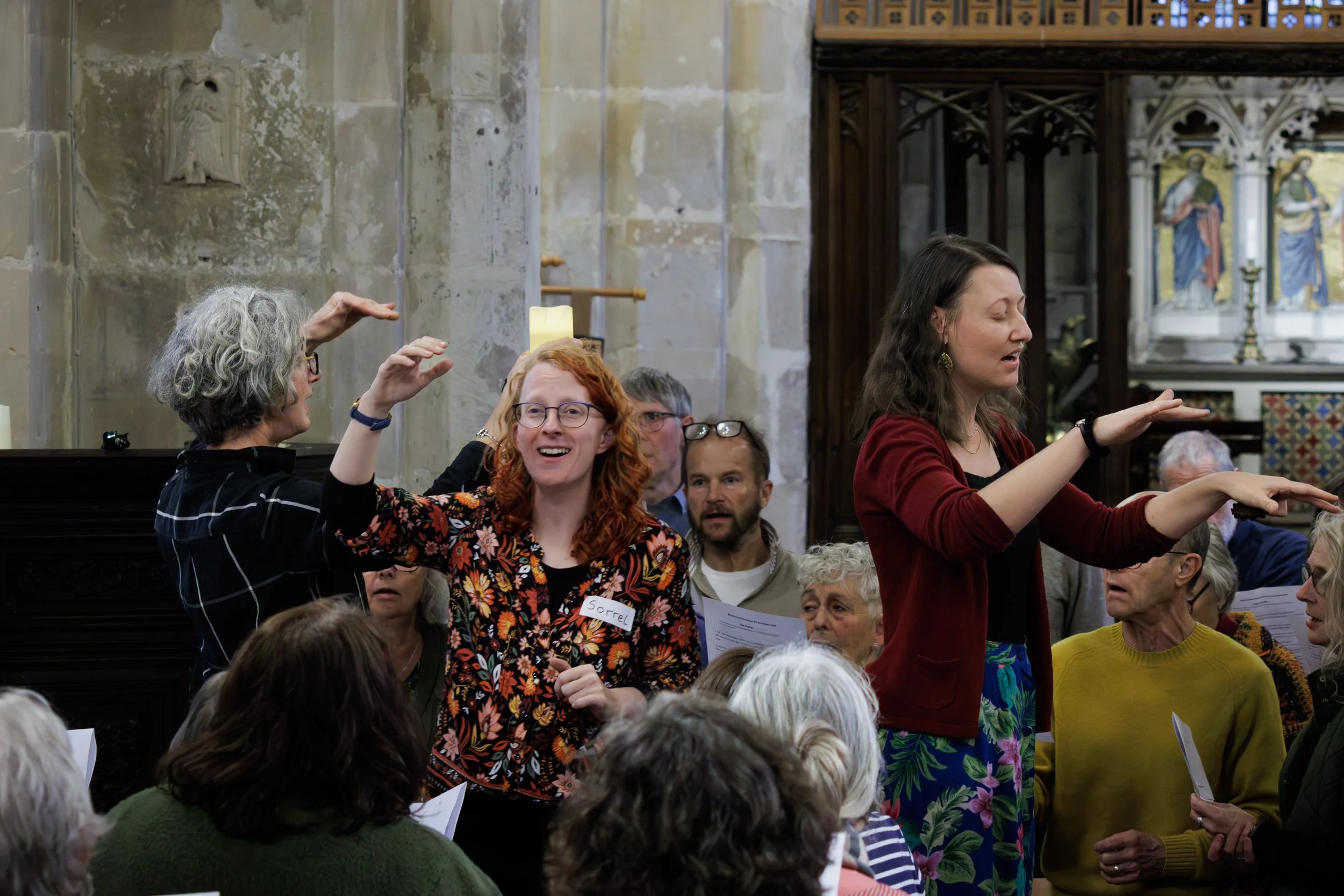 A group of people participating in a choir or singing event inside a church. Two women are standing with arms raised as if conducting or leading singing, while others are gathered around, holding sheets of paper. The background features historic church architecture with stained glass and stone walls.