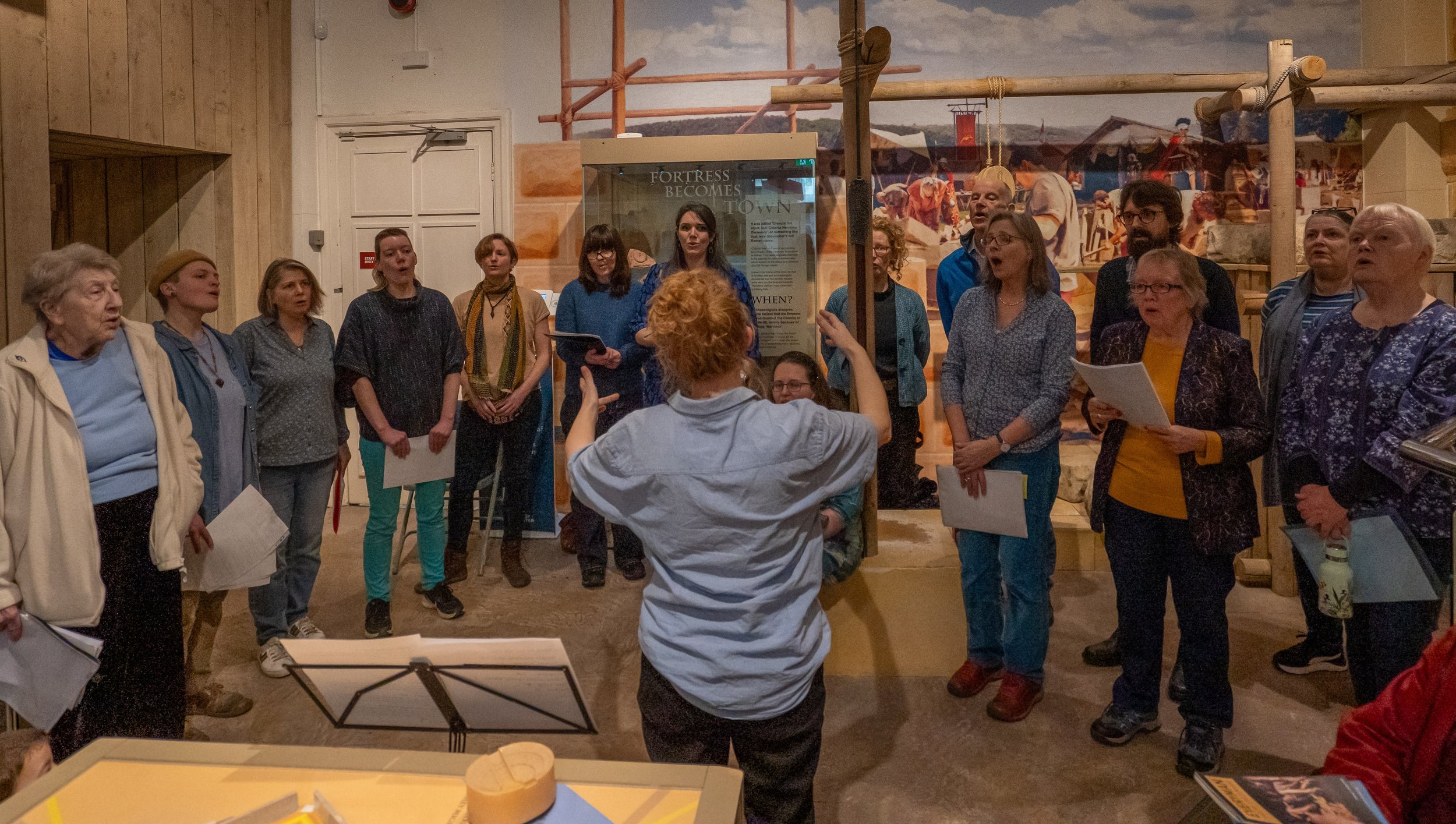 Group of people listening to a guide or tour leader in a museum or exhibit space. The guide is gesturing with her hands, and the group appears engaged. The background features a mural or display with historical scenes and a sign that reads 'Fortress Becomes Town.'