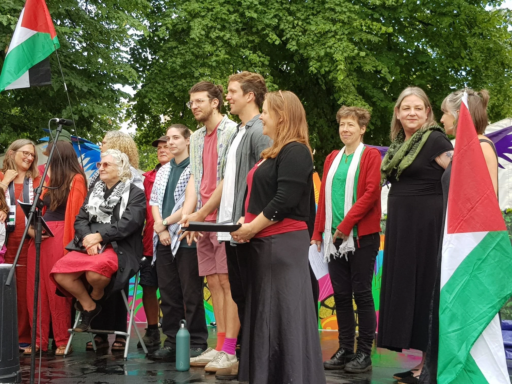Group of protesters standing outdoors in front of a colorful rainbow-themed banner during a demonstration, with green trees overhead and a flag on the left side of the image.