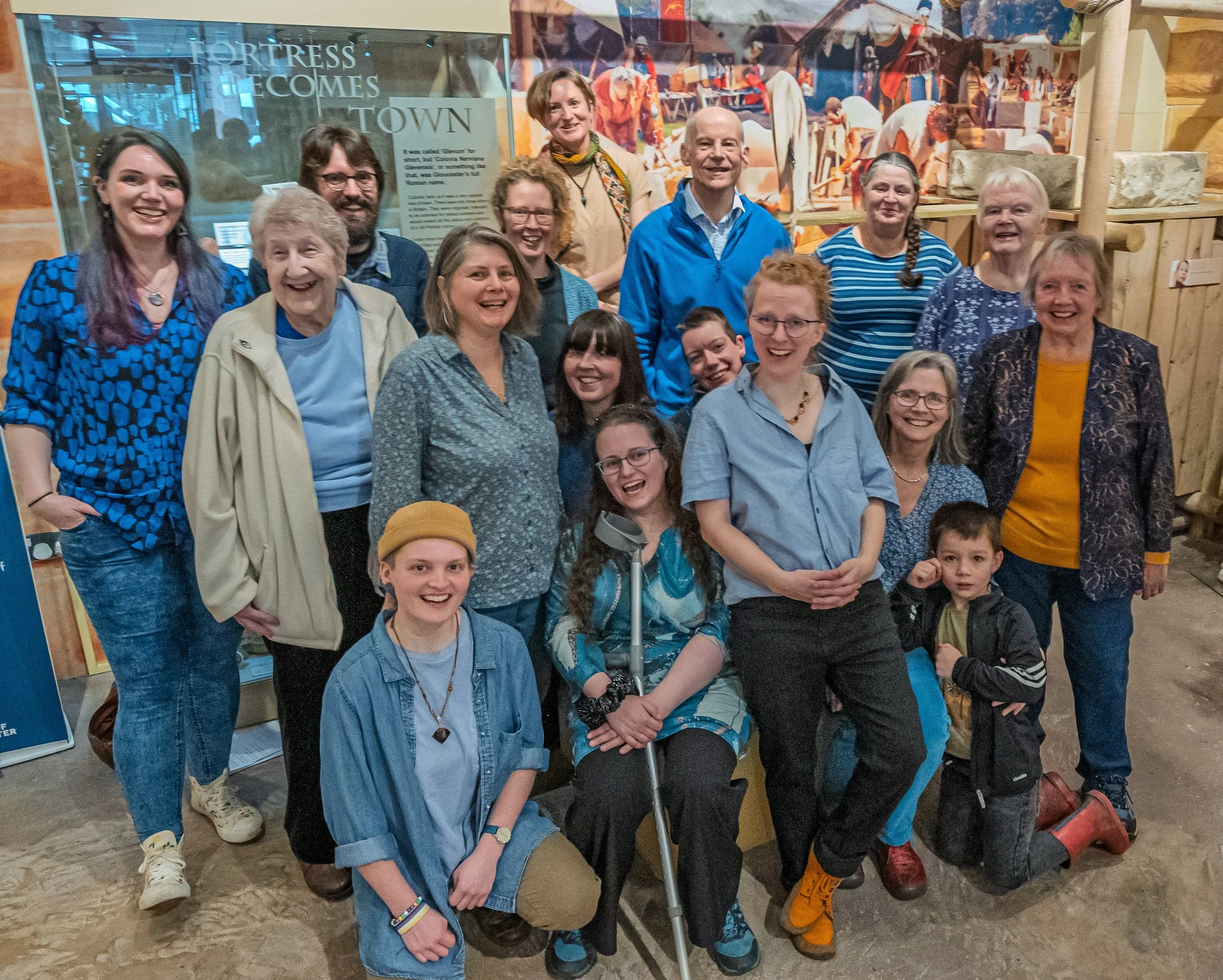 A diverse group of people, including children, adults, and elderly individuals, smiling and posing together in an indoor setting with a mural of historical or cultural scenes in the background.