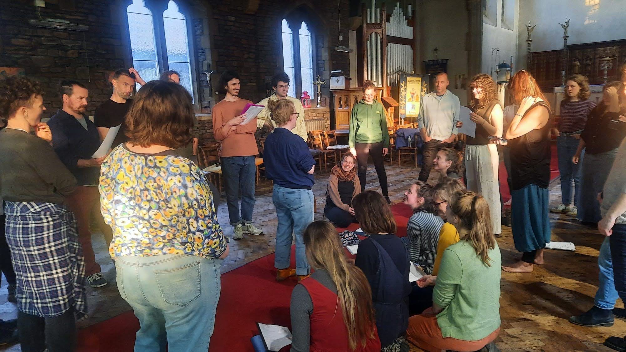 Group of people gathered inside a church, some standing and some sitting on the floor, participating in a religious or community event with a man at the center holding a paper and others listening attentively.