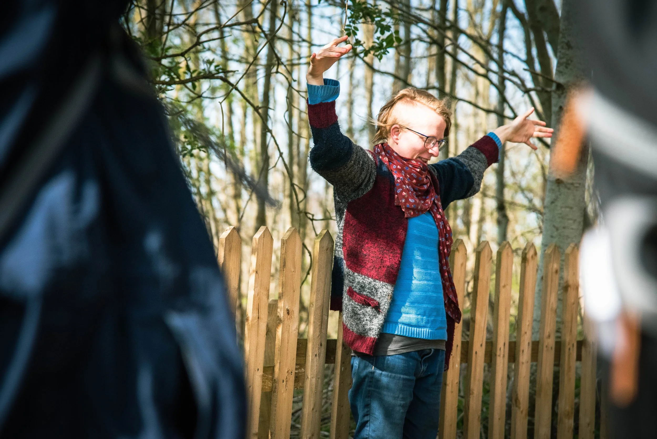 A woman with glasses, wearing a colorful sweater and red scarf, standing outdoors with her arms raised, in front of a wooden fence and trees.