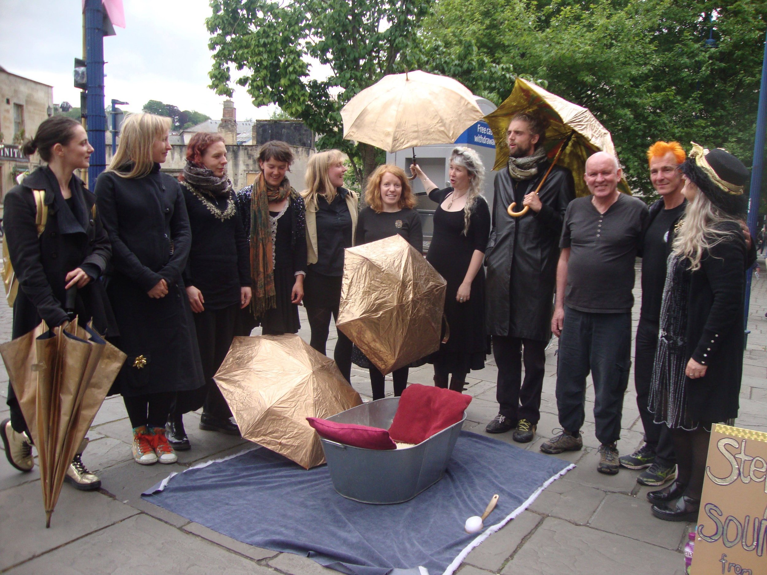 A group of people standing outdoors, some holding umbrellas, around a small display or puppet theater set-up with a metallic gold umbrella and fabric on the ground. Several appear to be engaged in conversation or amused by the scene.