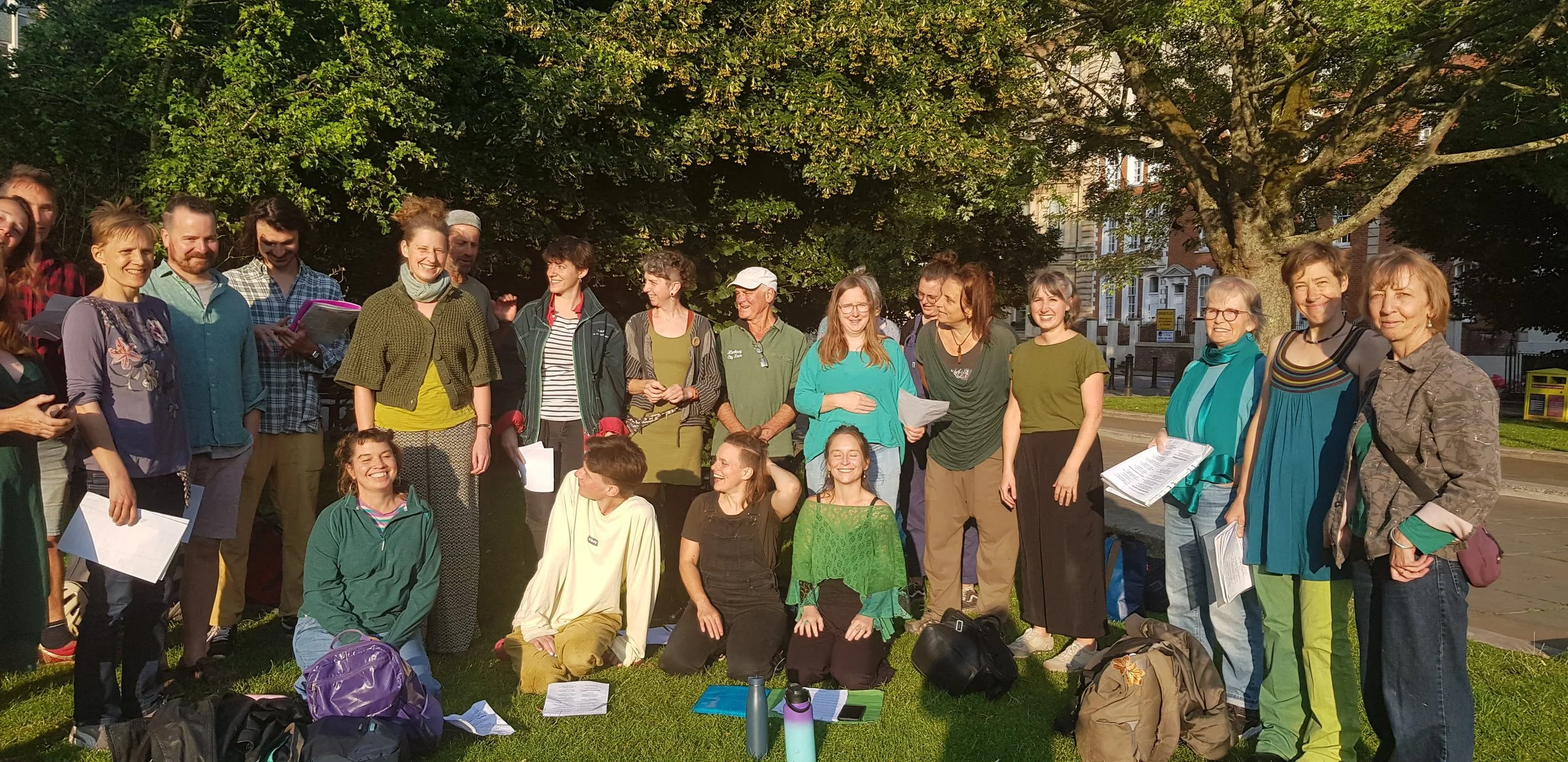 A group of people outdoors under a large tree, gathering and smiling for a photo. They are holding papers and backpacks, and some are sitting while others stand. It appears to be a sunny day.
