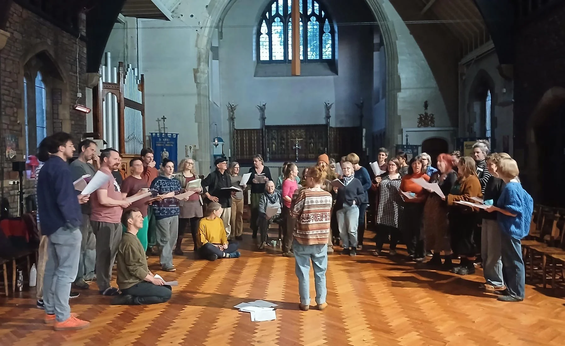 A group of people singing or rehearsing in a church with high arched ceilings and stained glass windows, some holding sheet music, with a person leading in the center, and others kneeling or standing around.