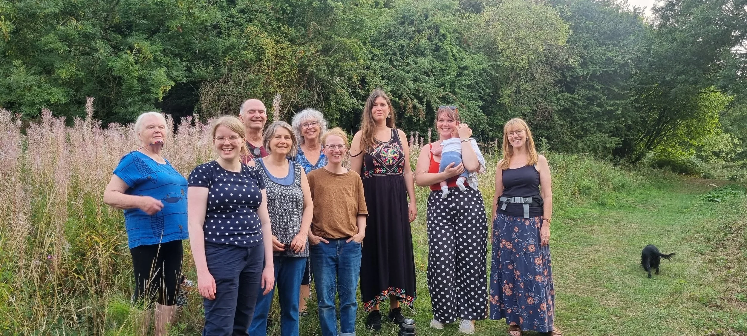 A group of ten people and one small black dog standing outdoors in a natural setting with greenery and trees in the background. The group includes men and women of various ages, dressed casually, some smiling and relaxed.