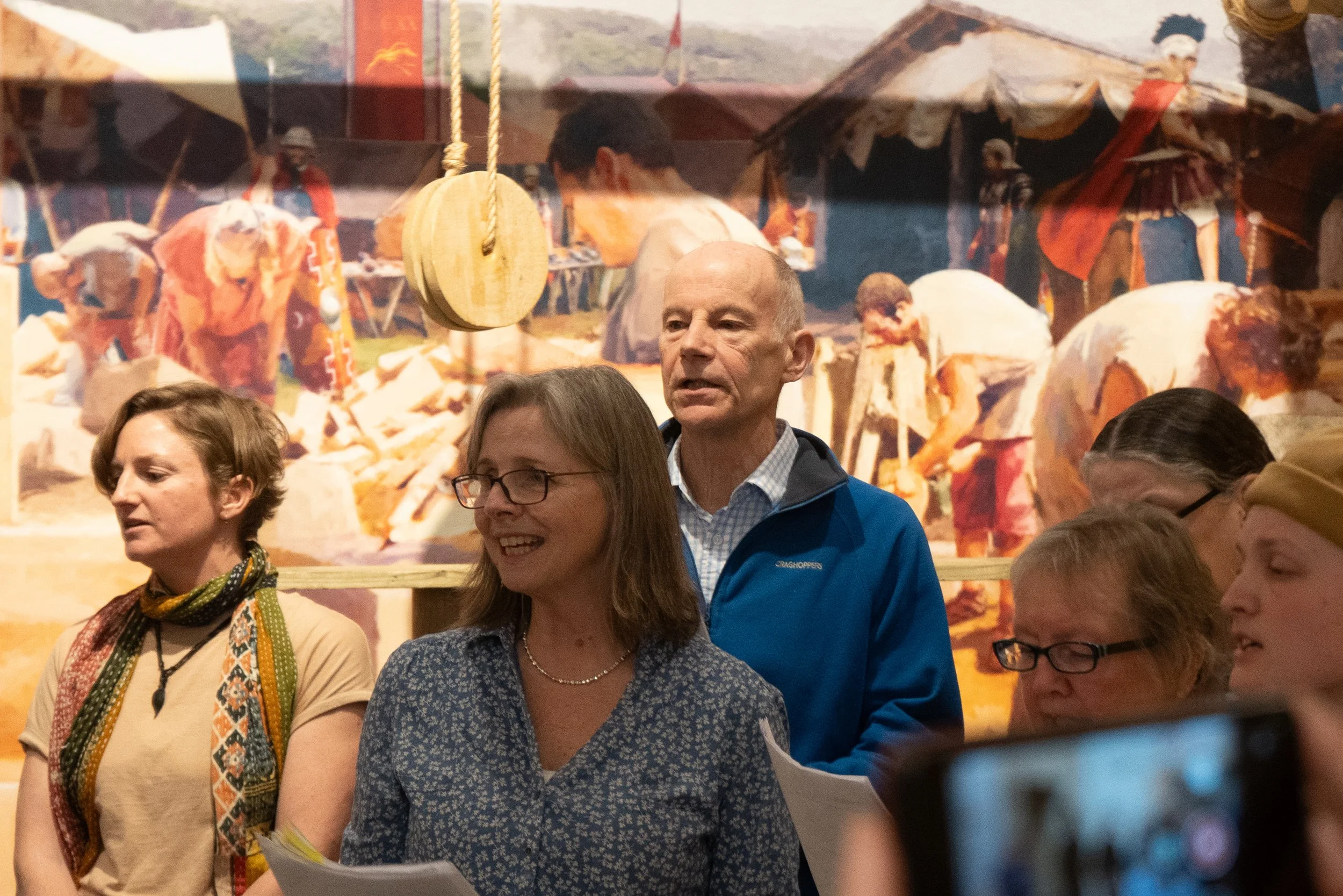 Group of people standing indoors in front of a colorful backdrop depicting a market scene with vendors and sheep.