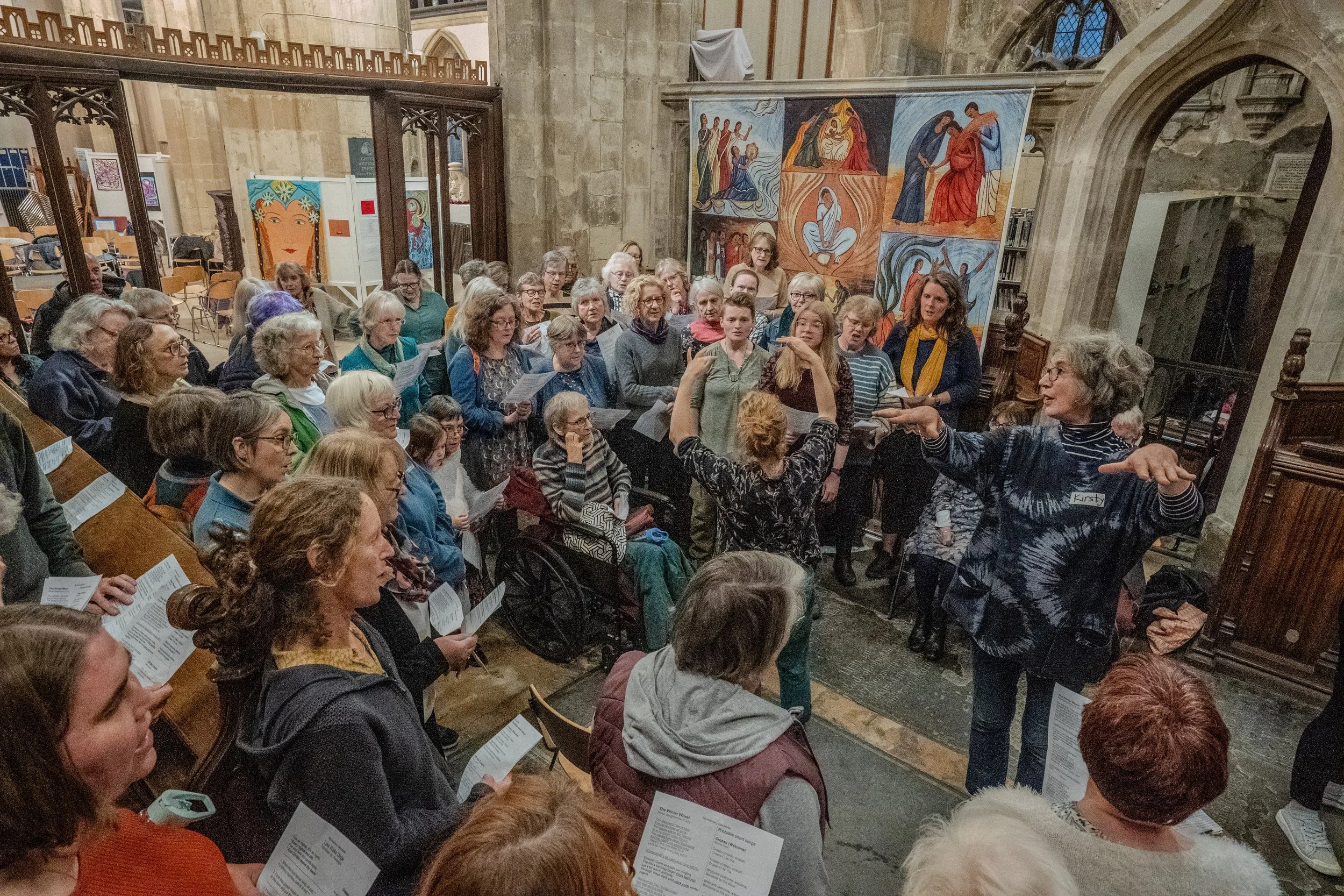 A large group of people gathered inside a historic church, participating in a choir or singing event, with church artwork and stained glass windows visible in the background.