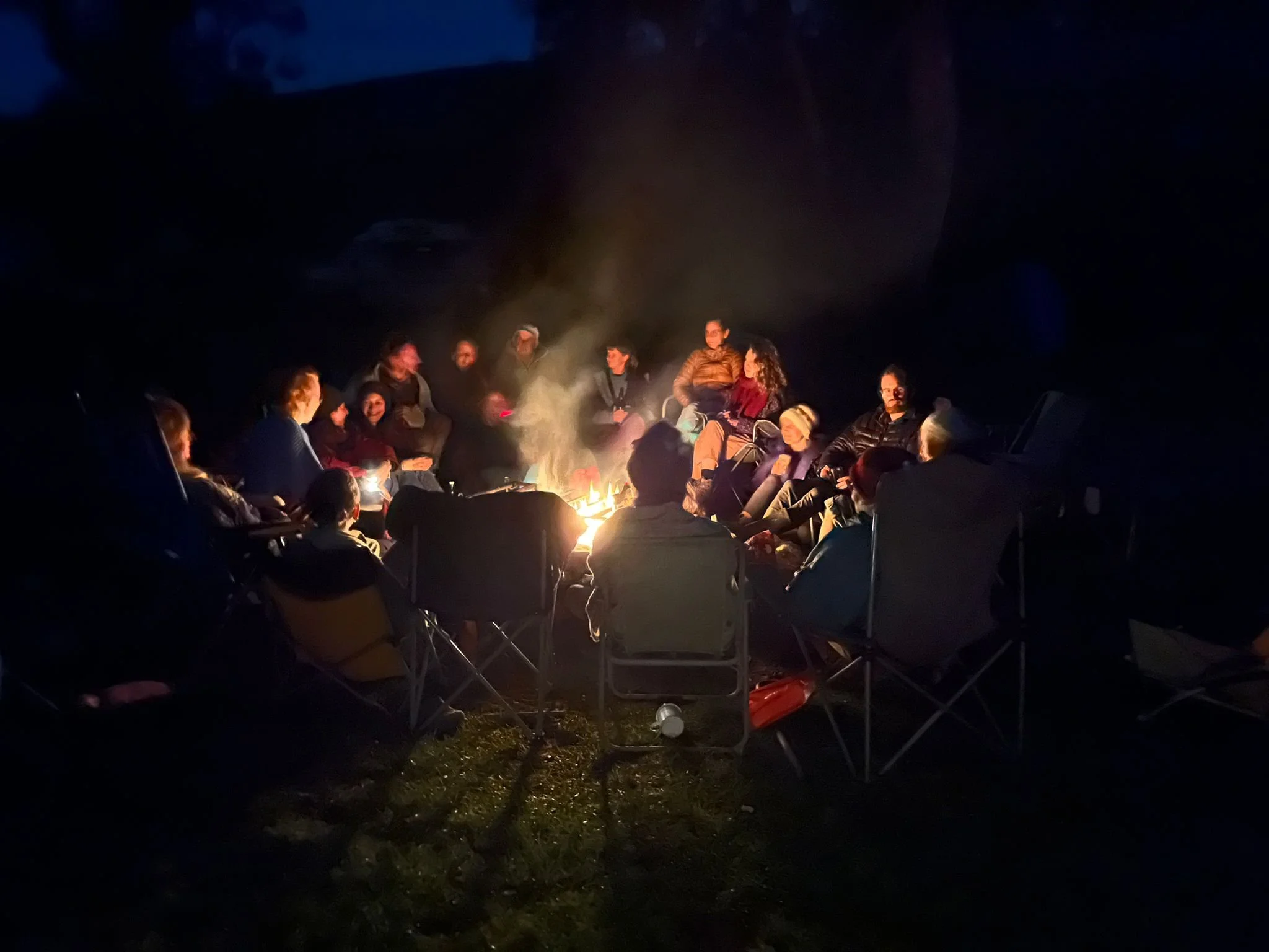 Group of people sitting around a campfire at night, some sitting on chairs, enjoying conversation and warmth.