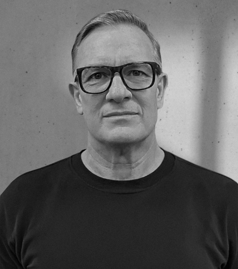 Black and white photo of a middle-aged man wearing glasses and a dark shirt, standing against a plain background.