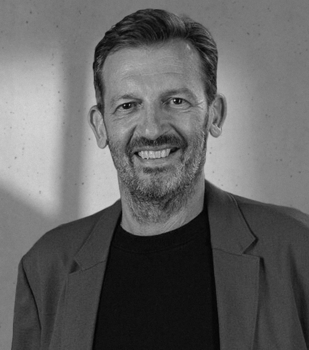 Black and white photo of smiling man with short hair, beard, wearing a blazer and dark shirt.