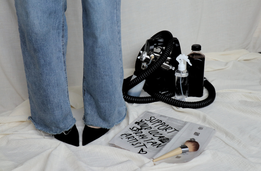 A person standing on a bed wearing jeans and black shoes, surrounded by cleaning supplies and a makeup brush, with a handwritten note saying 'Support Your Local Stay Team' and a few other items.