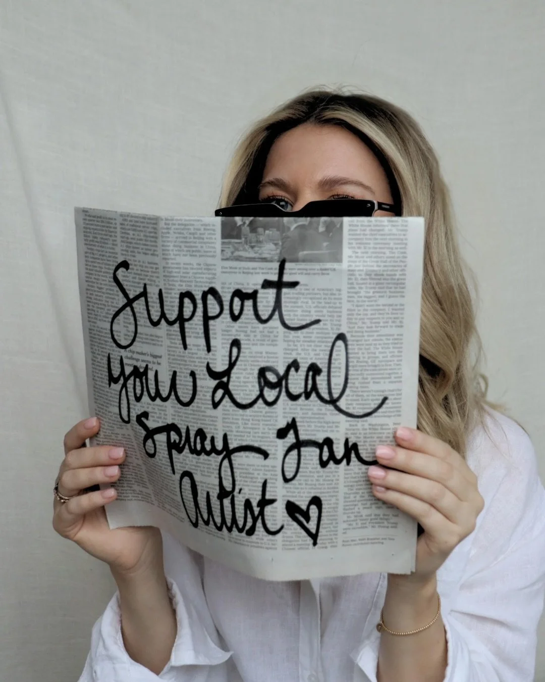 Woman holding a newspaper with a handwritten message supporting local charities, partially covering her face, against a plain background.