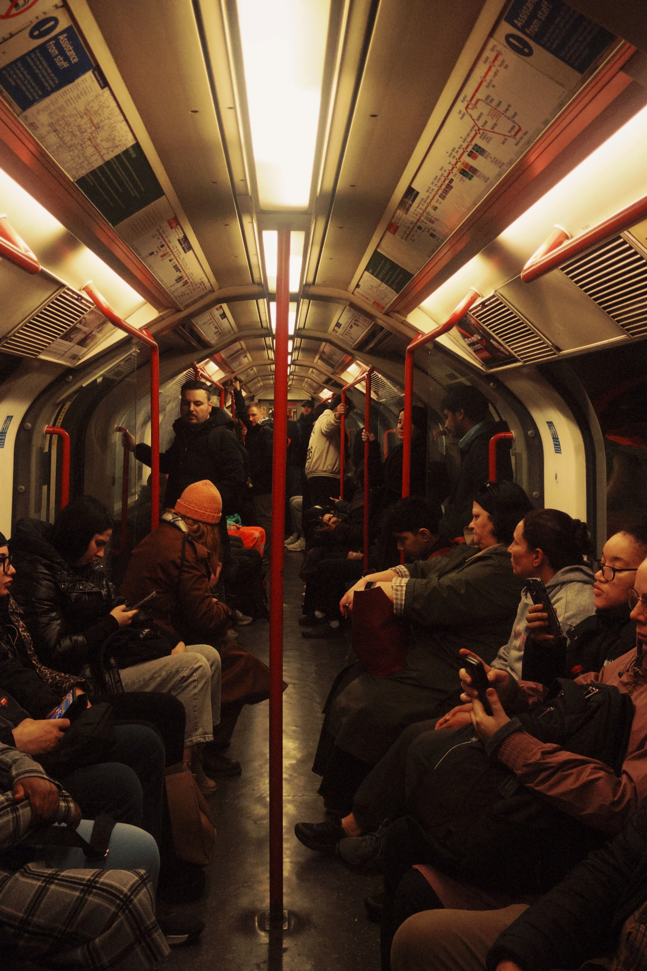 People sitting inside a subway train car, some looking at their phones, with overhead maps and red handrails visible.