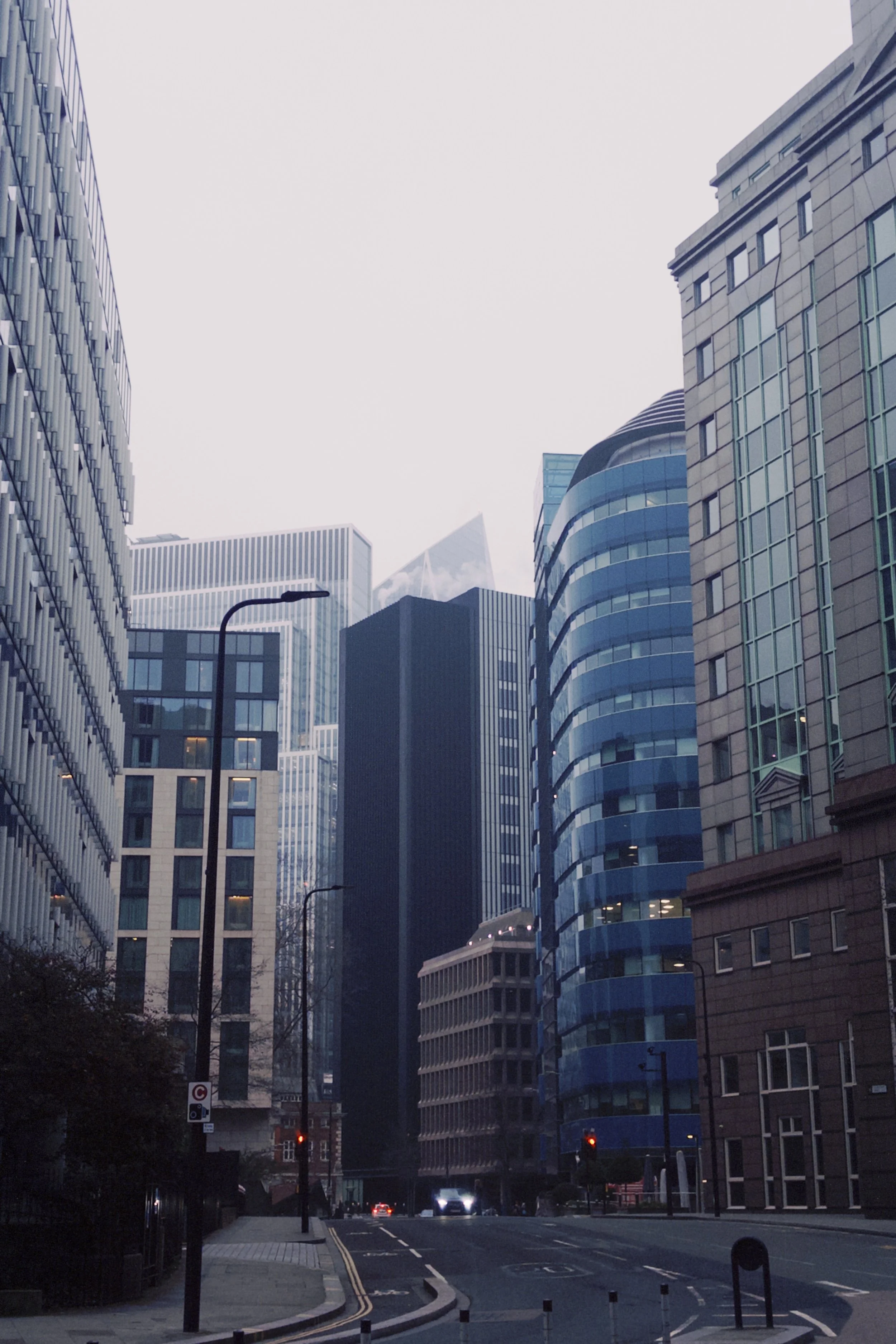 City street with tall modern office buildings on both sides, streetlights, cars, and a crosswalk, with overcast sky in the background.