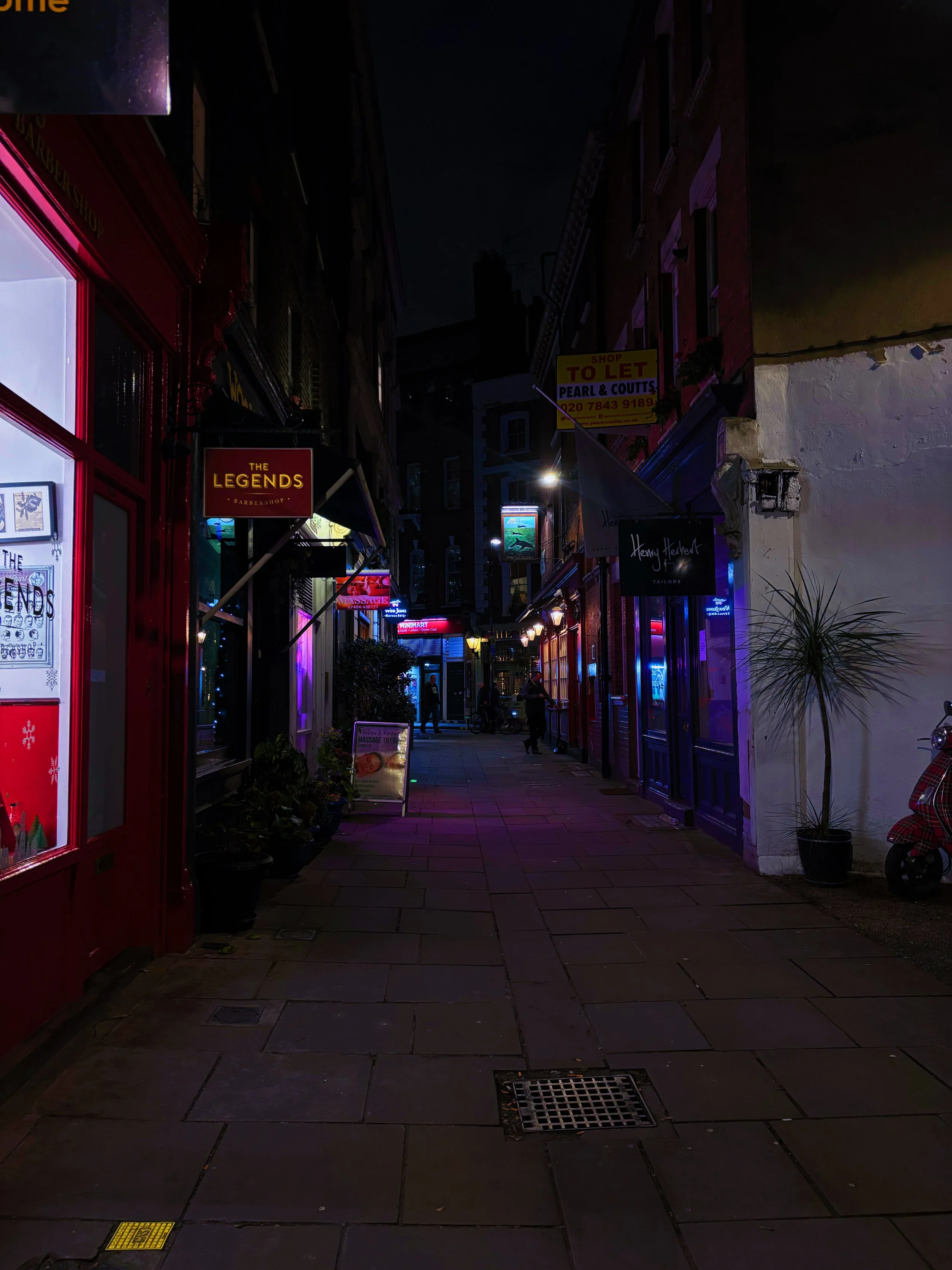 Nighttime view of a narrow street with various shop signs illuminated, including a barbershop sign and a sign for real estate to let, with a few people visible in the distance.
