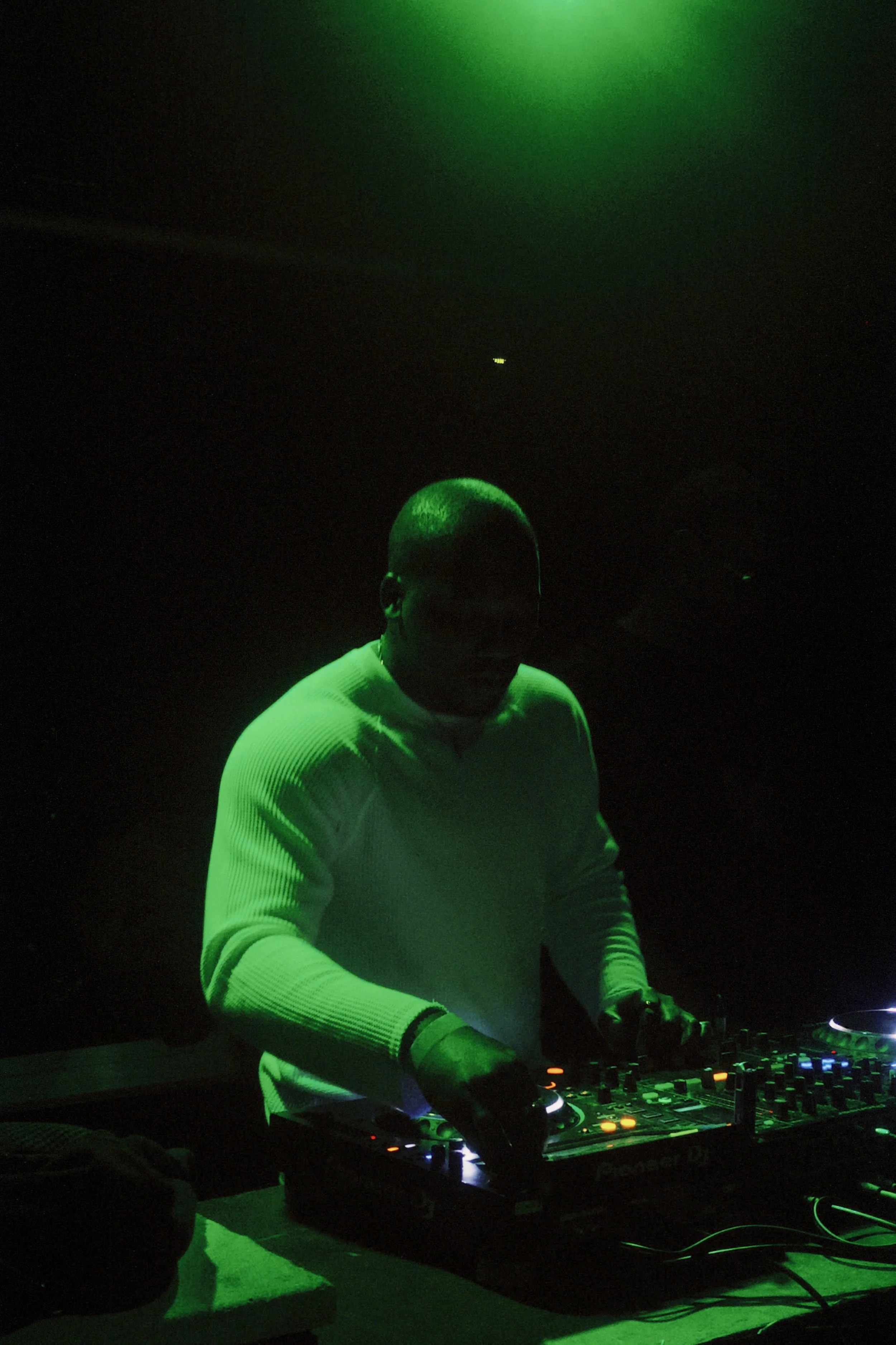 A DJ wearing a white sweater and headphones is performing with green lighting in the background, using DJ equipment.
