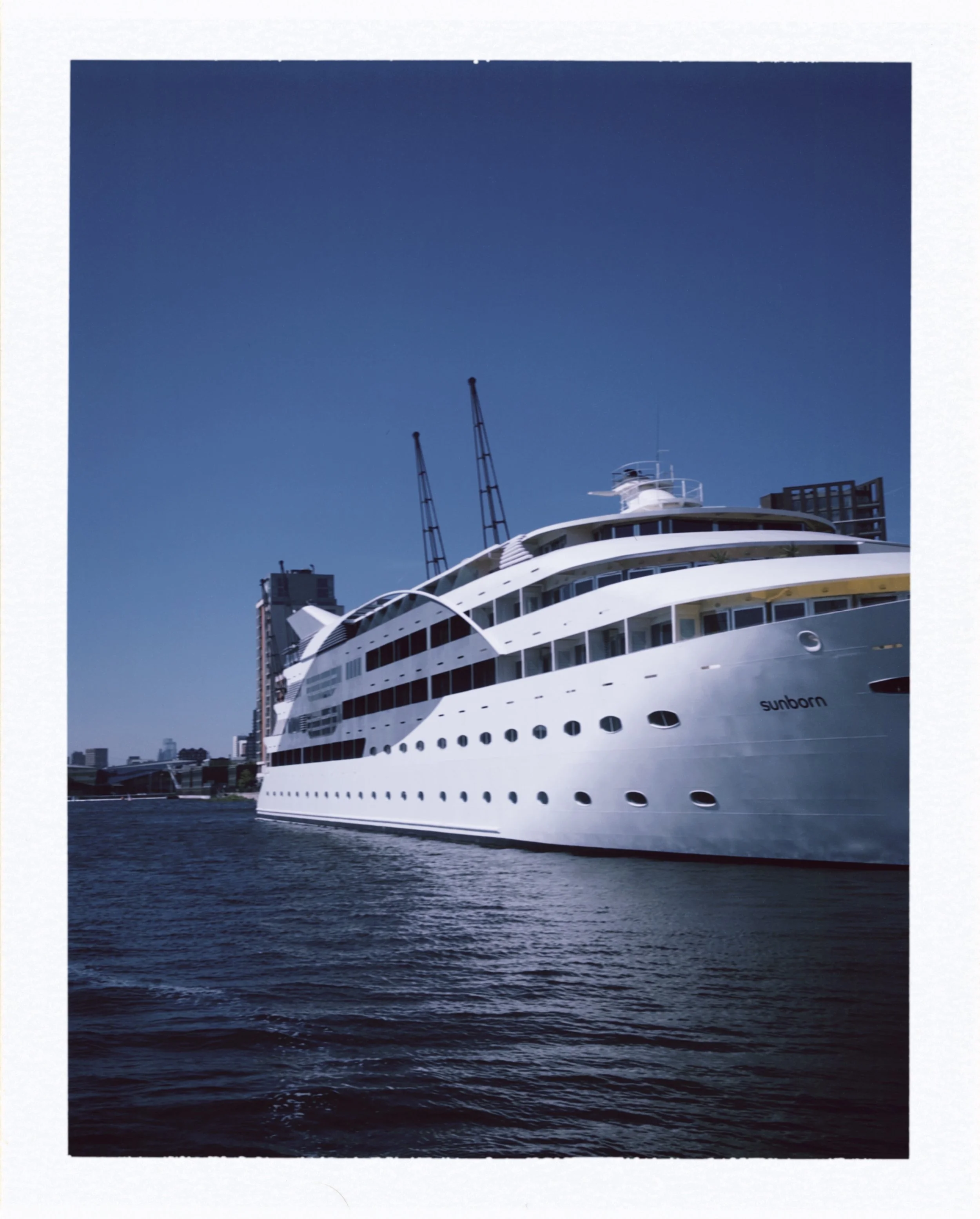 A large white luxury yacht named Sunborm docked on the water with a cityscape and tall cranes in the background, under a clear blue sky.