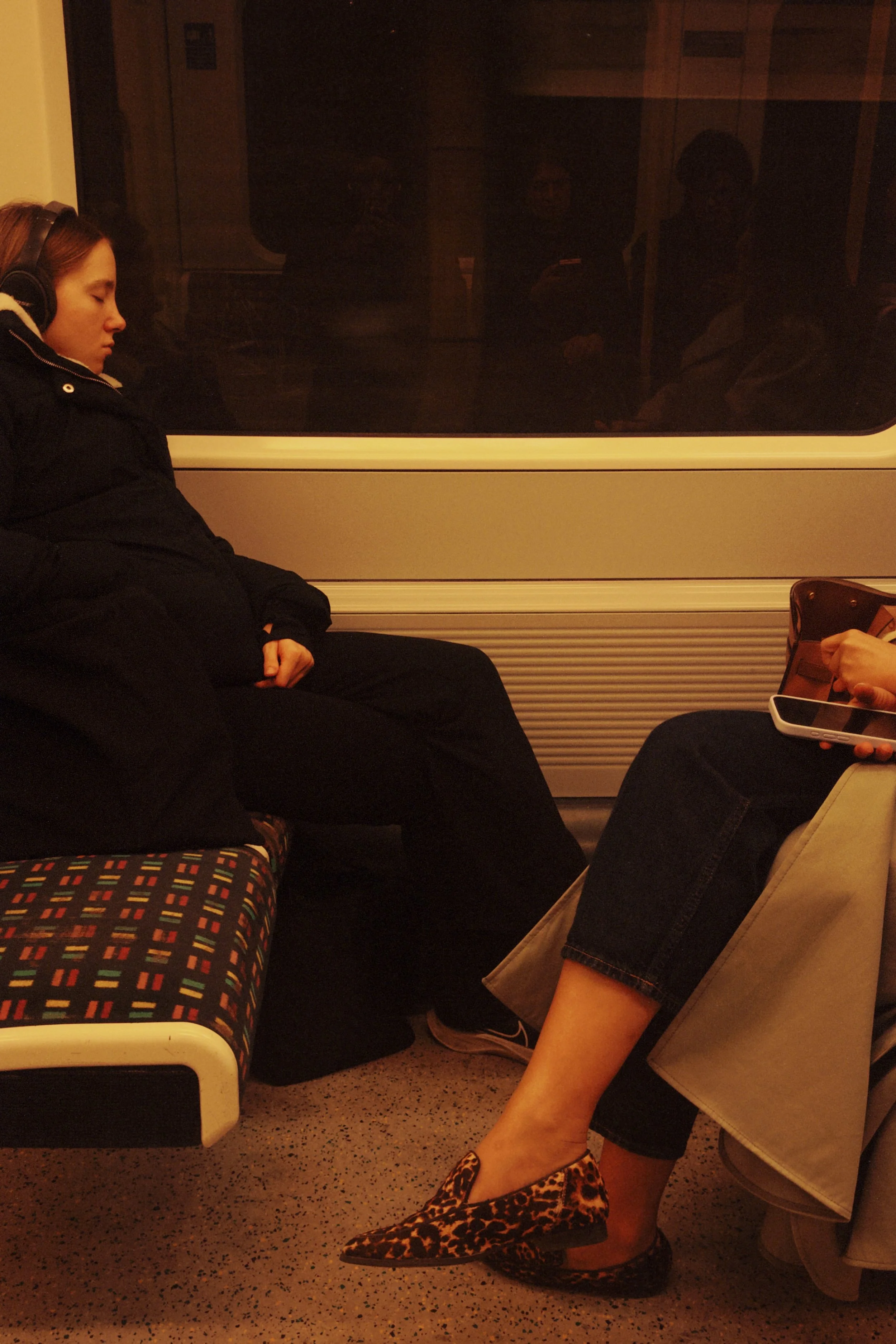 A woman with leopard print shoes sitting with her legs crossed on a subway train. In the background, other passengers are visible through the window, some standing and some using their phones.