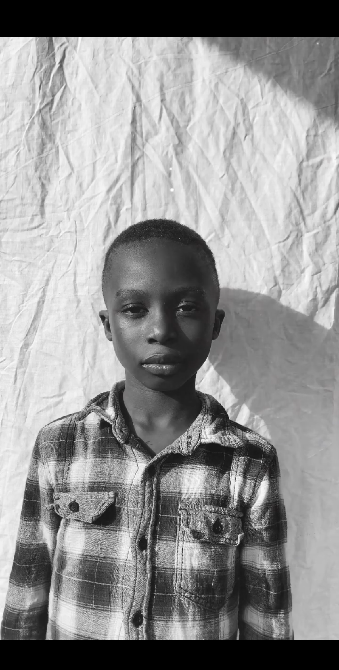 A young boy with short hair, wearing a plaid button-up shirt, stands in front of a crinkled fabric backdrop.