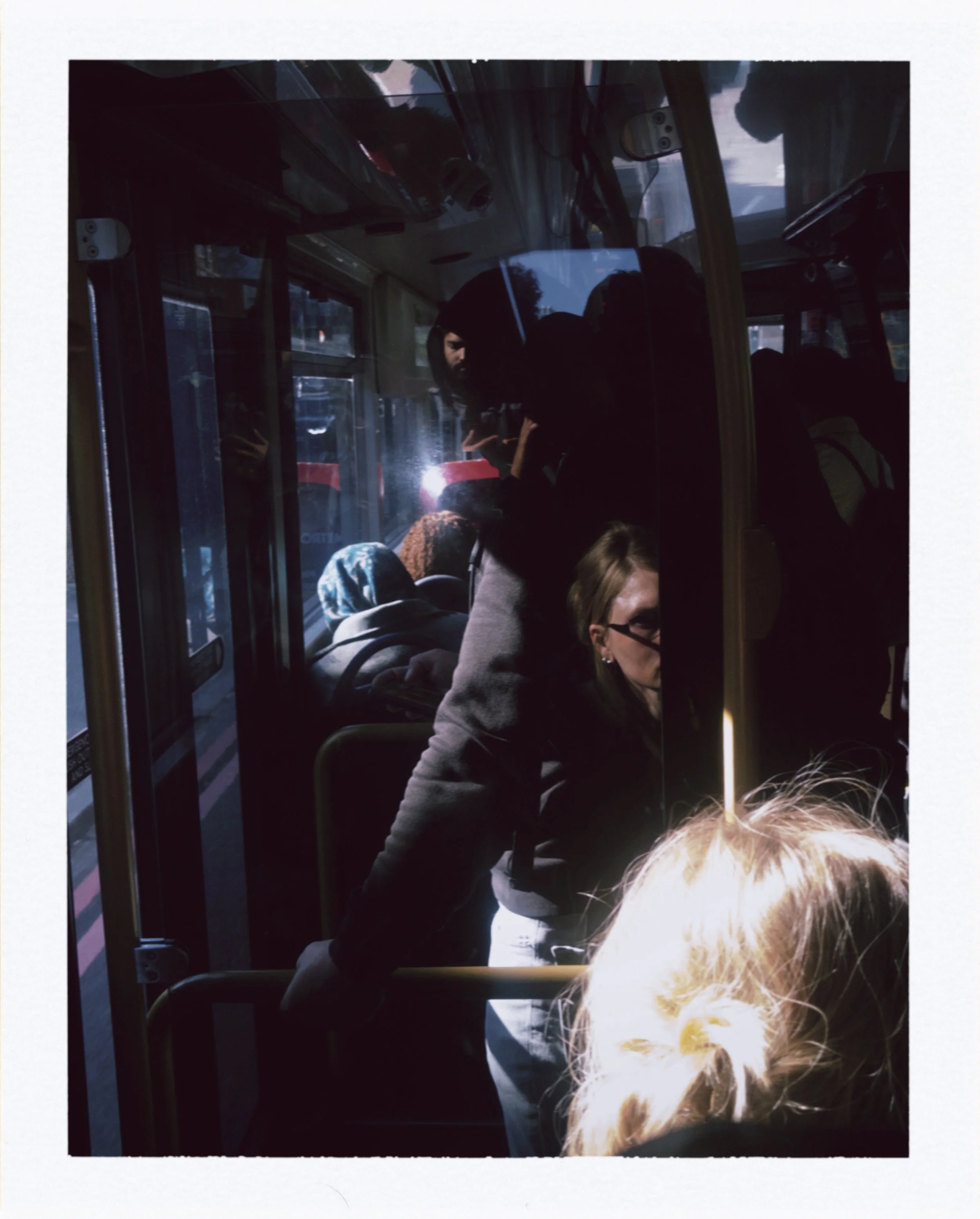 People sitting on a bus, some visible through the window, with natural light coming in, and one person in the foreground with light-colored hair.