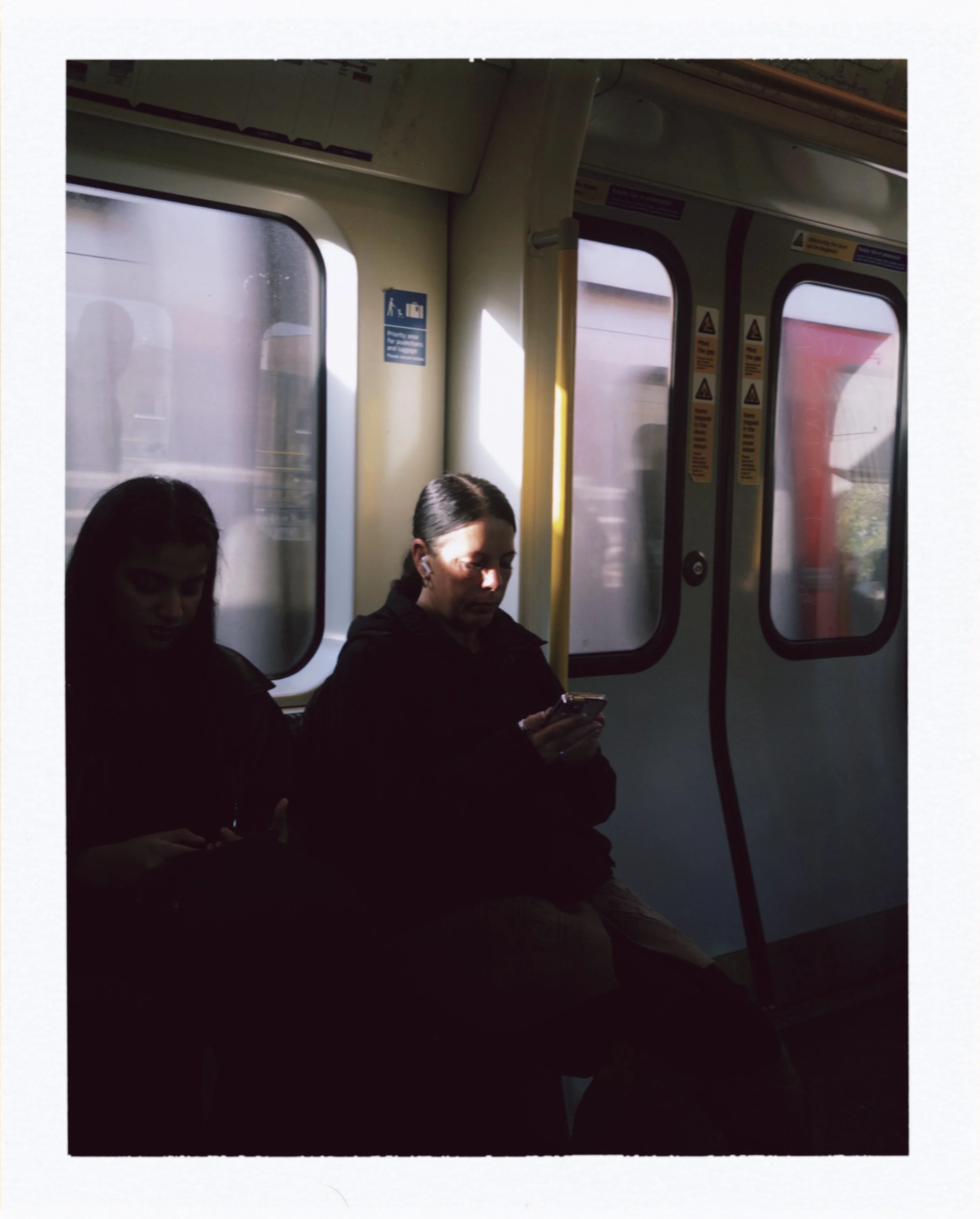 Inside of a train with two women looking at their phones, windows showing a blurred outdoor scene, and safety and instruction signs on the walls.