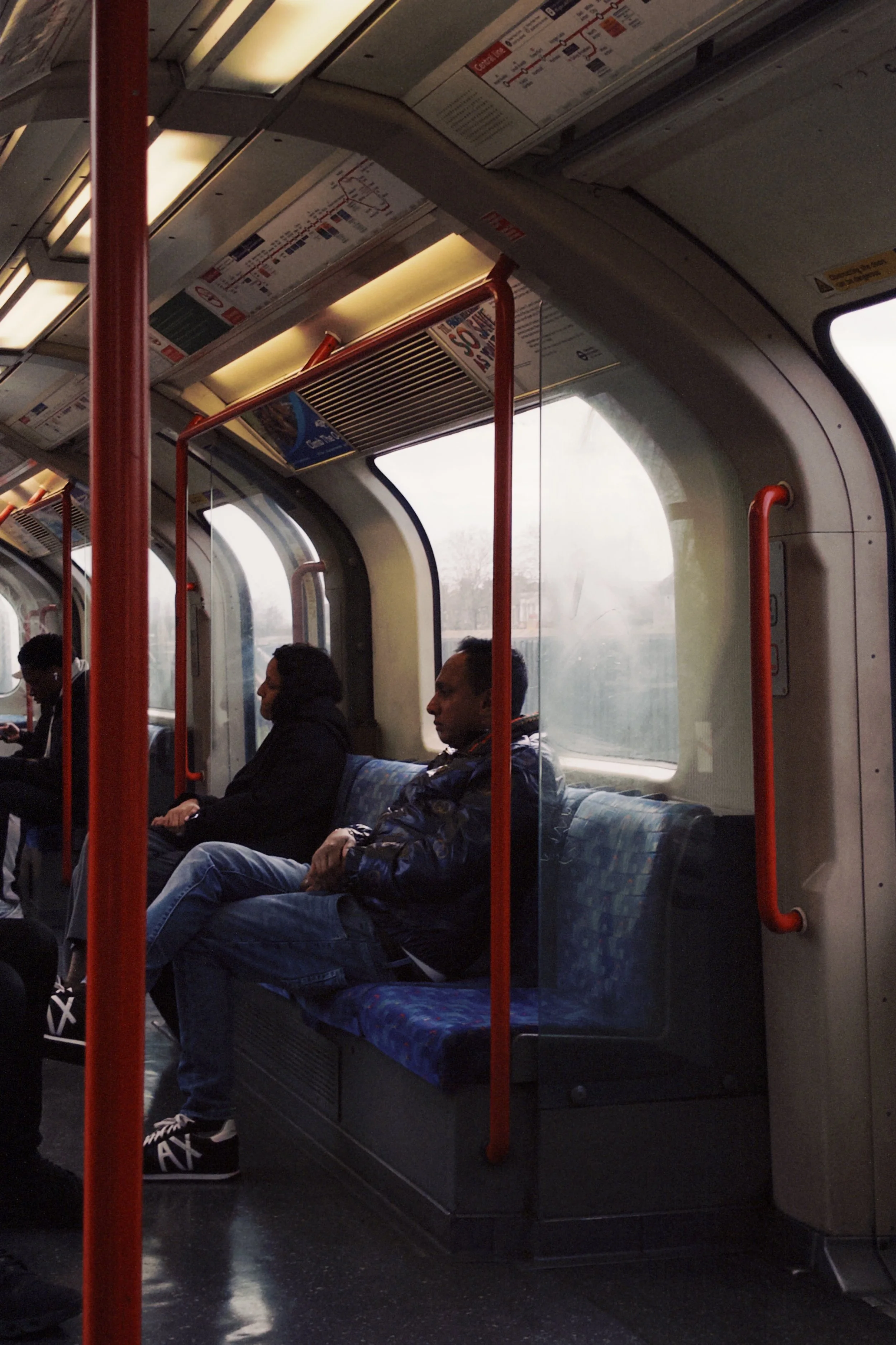 Inside a bus with passengers sitting on blue patterned seats, red handrails, large windows showing a cloudy outdoor scene, and overhead advertisements.