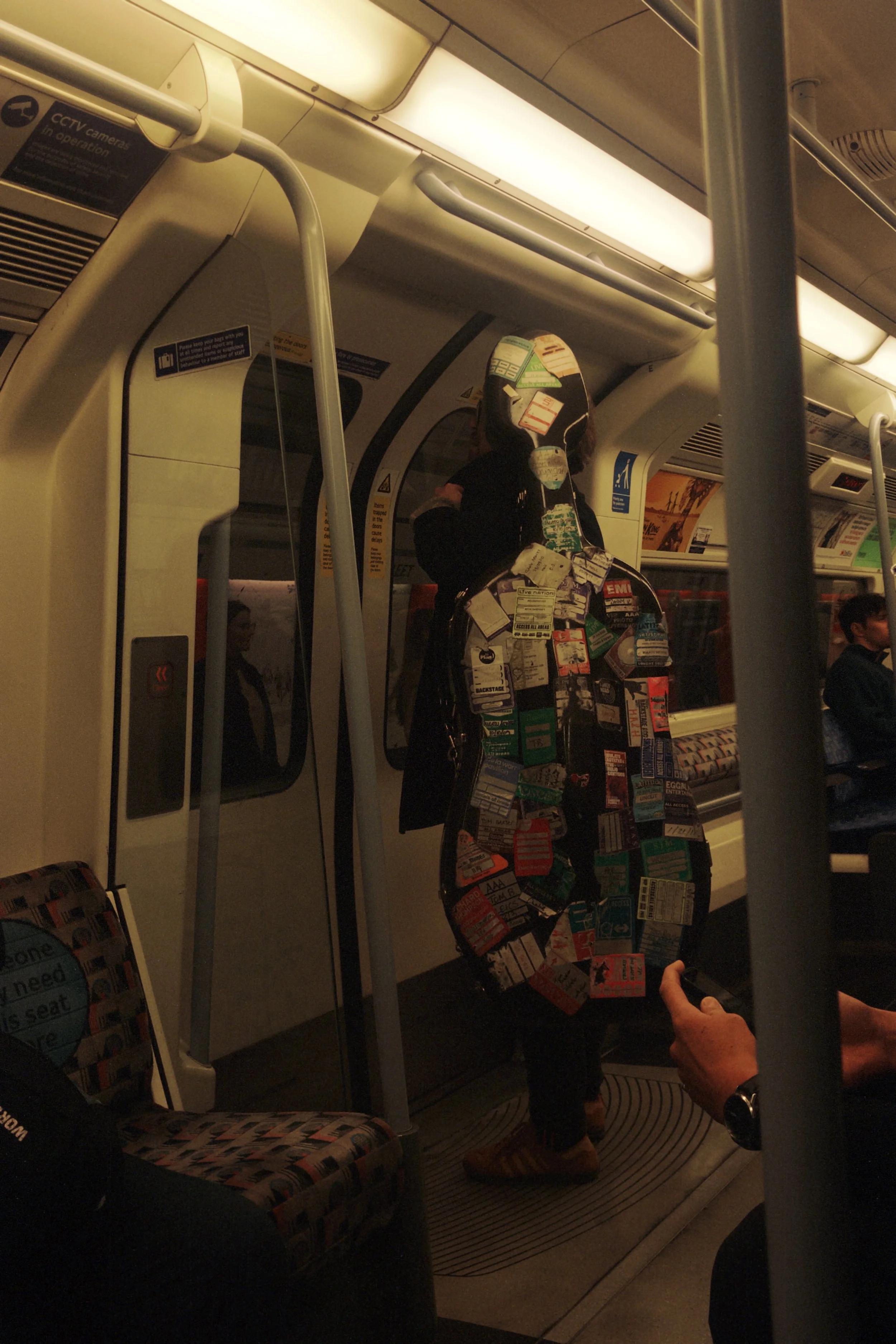 Inside a subway train, a person stands holding onto a handle, their body covered in tickets and labels. Other passengers are seated or standing in the background, and the subway door is visible.