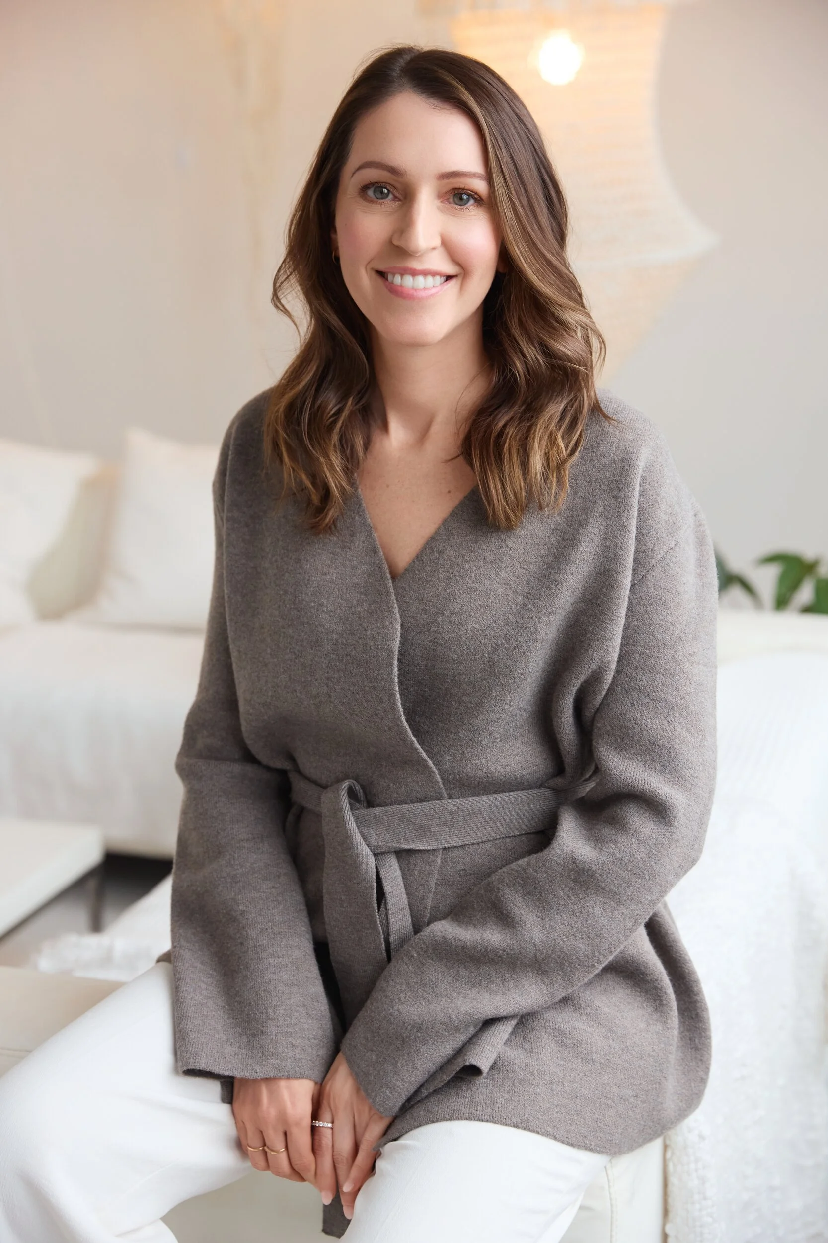 Une femme souriante avec des cheveux bruns longs et ondulés, portant un manteau gris et un pantalon blanc, assise dans un intérieur lumineux avec un lampadaire en arrière-plan.