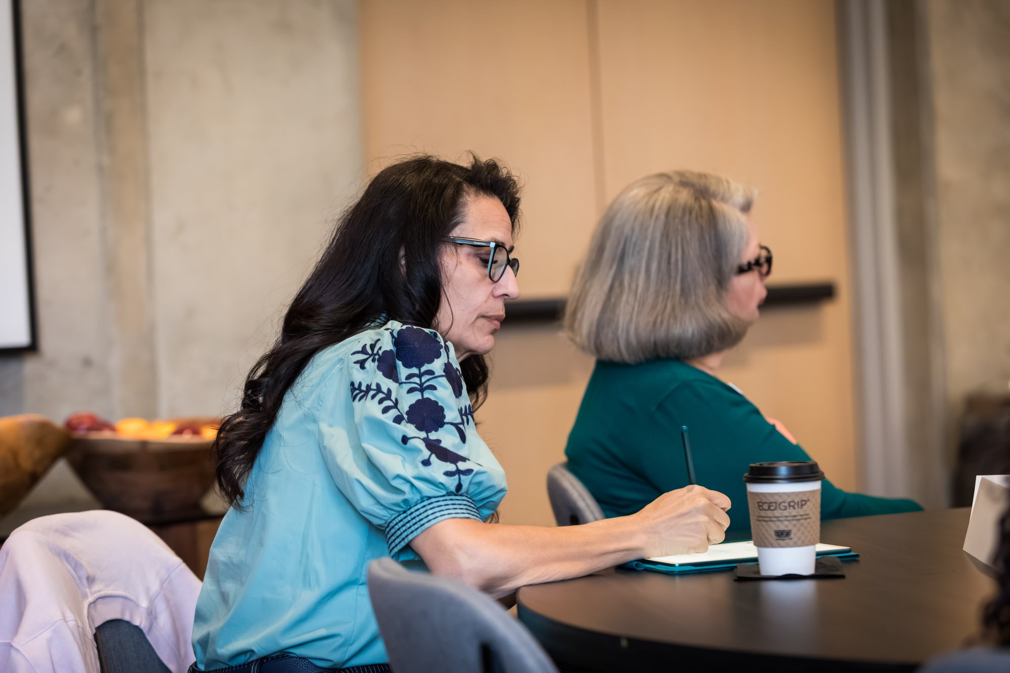 Two women sit at a table; one takes notes on a notepad, and a coffee cup is on the table in front of her looking over the FFTA research approach.