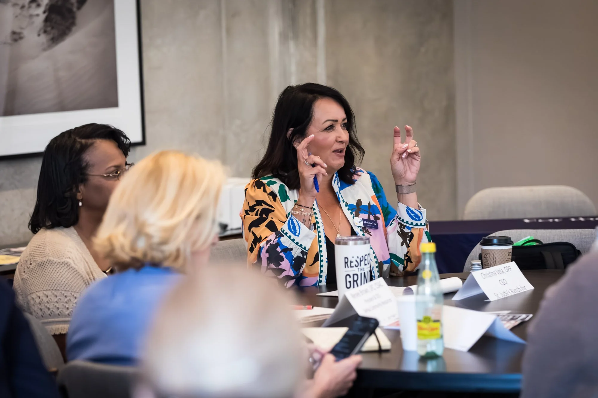 Woman in patterned jacket speaks about the child welfare policyand gestures with air quotes during a meeting, seated with others at a conference table.