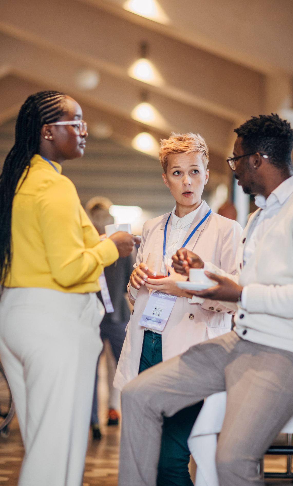 Three people wearing conference badges converse while holding drinks in a well-lit indoor setting.