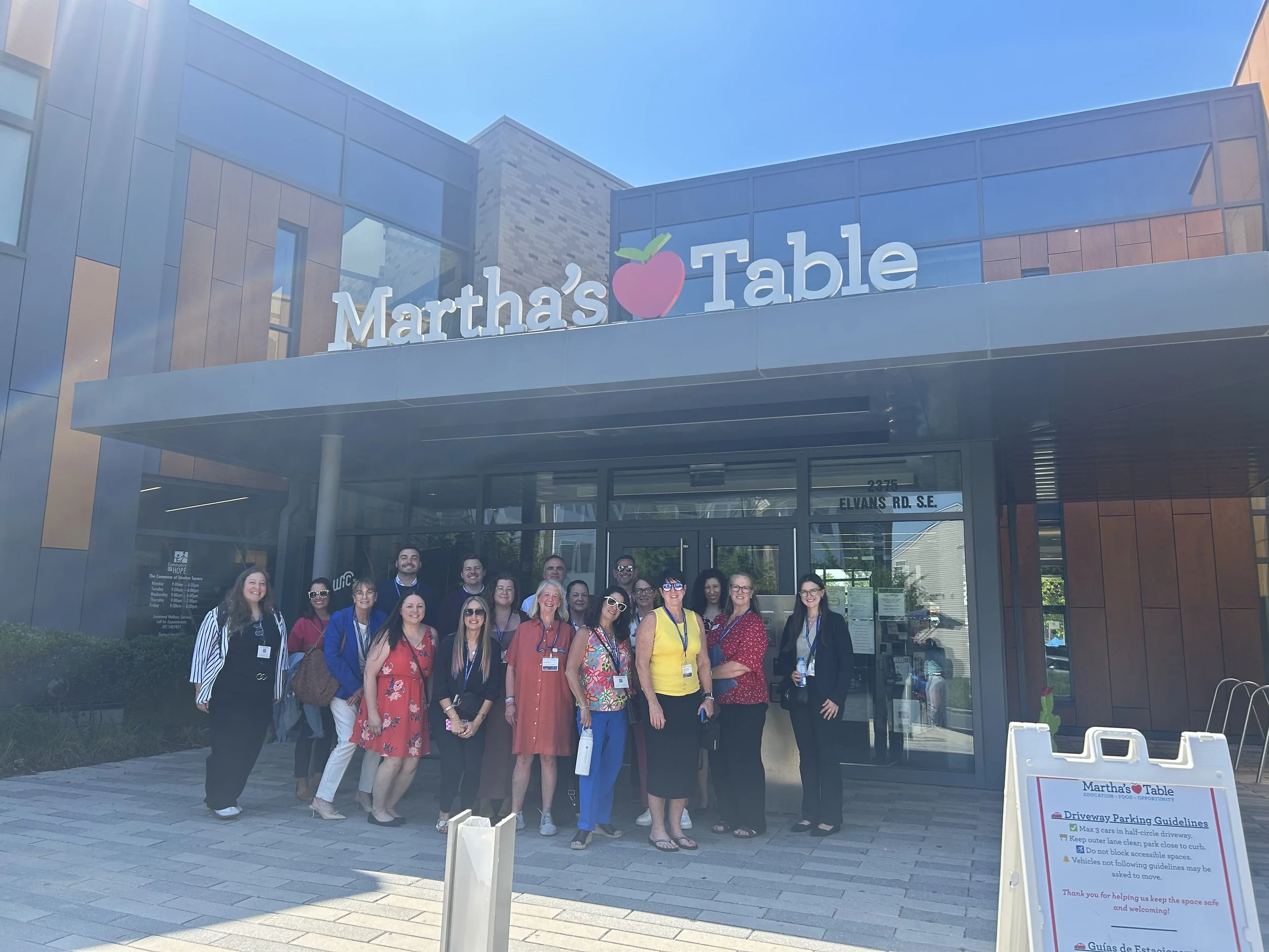 A group of FFTA community stands in front of the entrance to Martha’s Table, a community organization, on a sunny day.