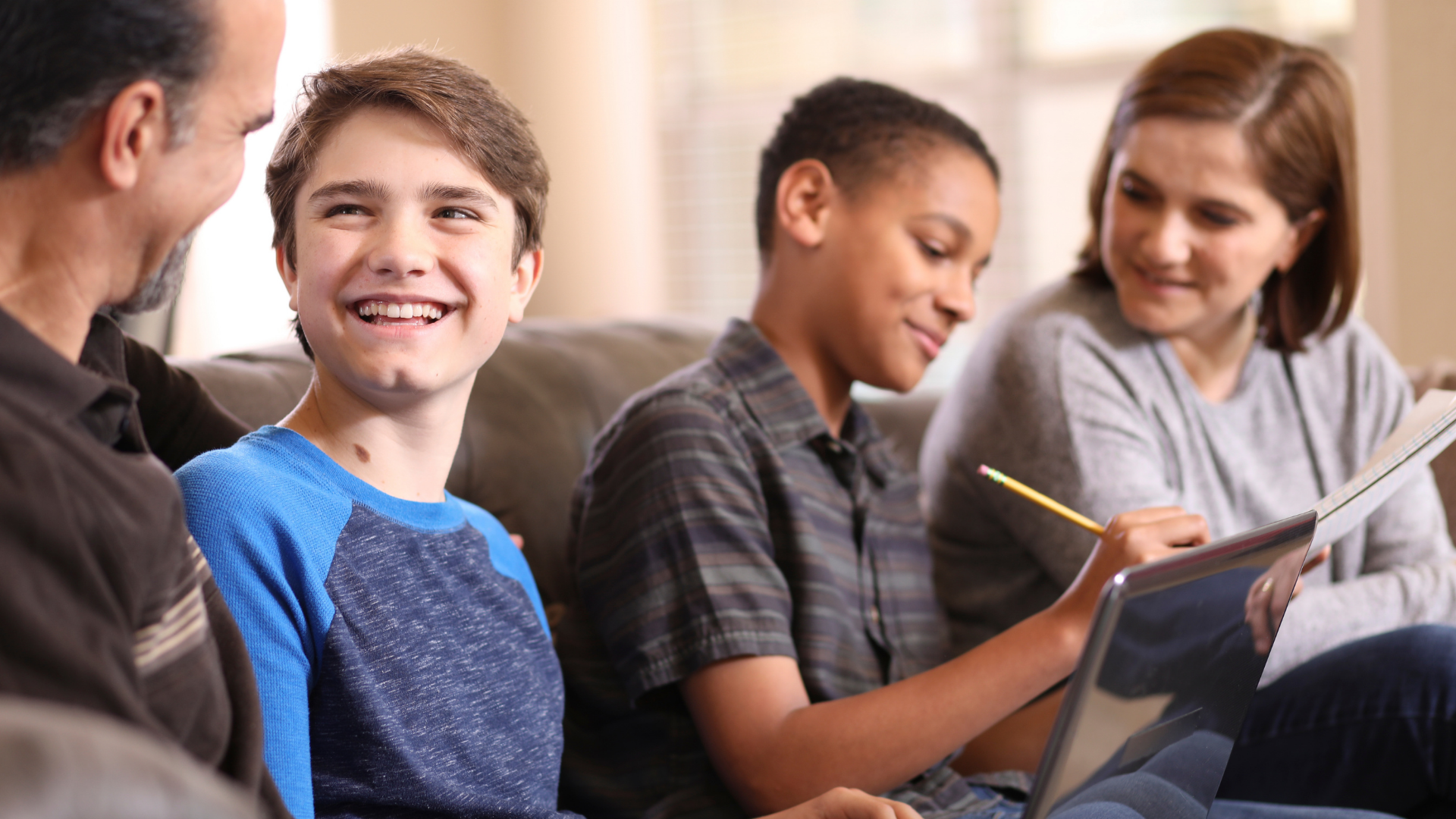Group of four diverse people sitting on a couch, smiling and engaging in conversation, with two of them using tablets.