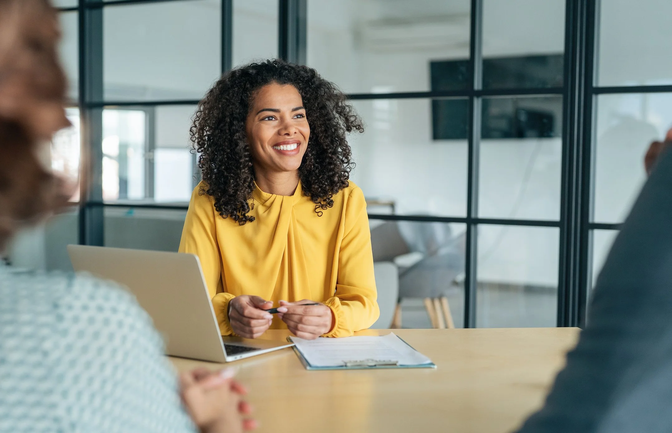 Woman in yellow blouse sits at a desk with a laptop and clipboard, smiling during a meeting about family well-being organizations.