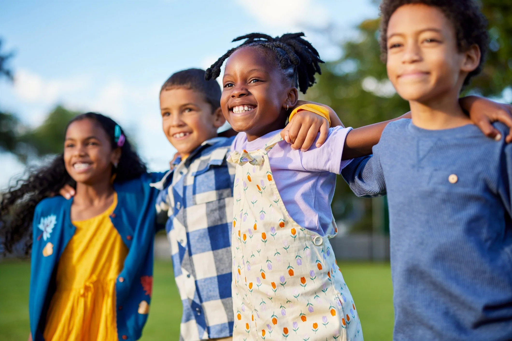 A group of diverse children with their arms around each other's shoulders outdoors, smiling and enjoying a sunny day.
